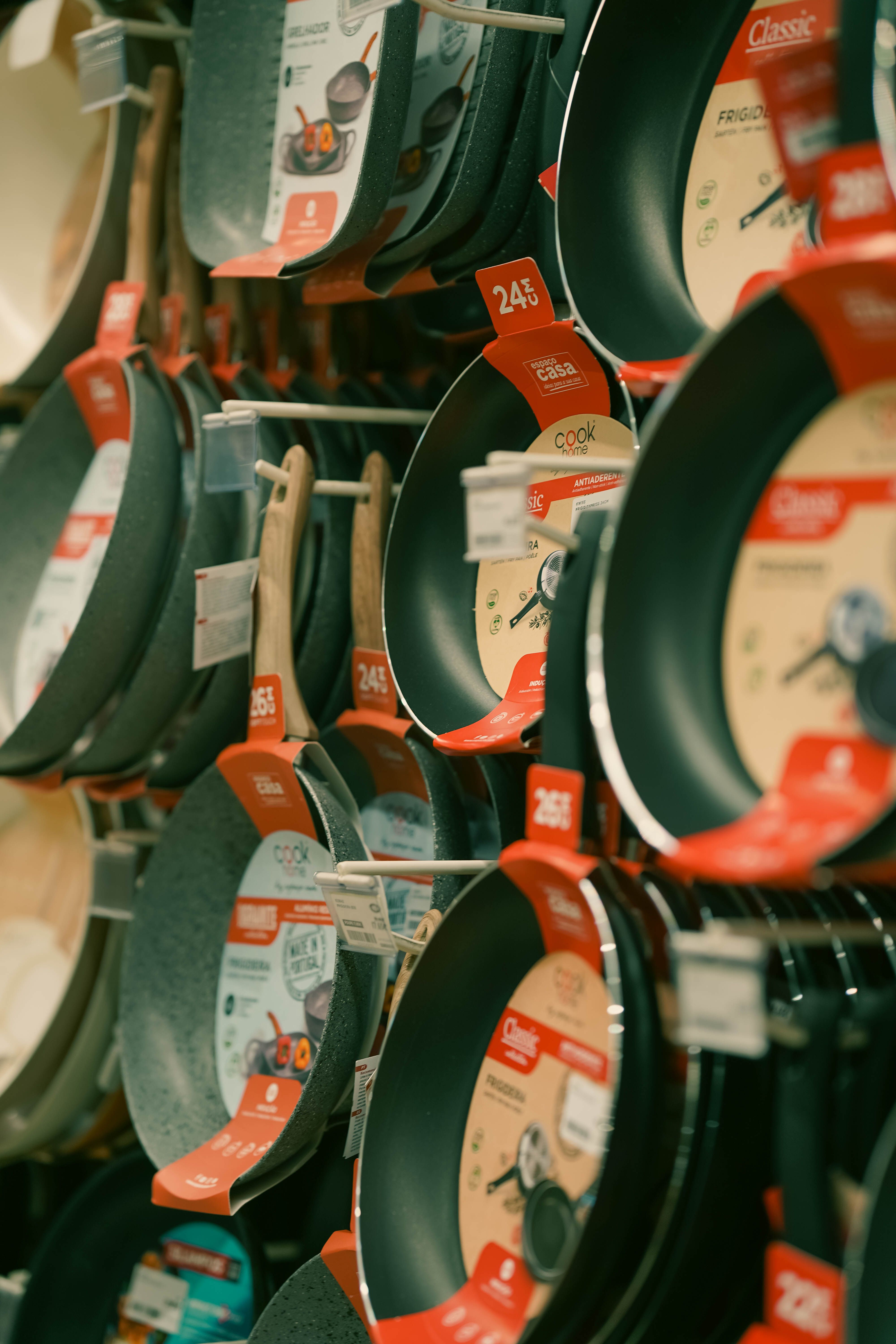 Dans un magasin d'ustensiles de cuisine, un présentoir mural où sont accrochées, sur des tringles, des poêles antiadhésives de différentes tailles, chacune munie d'une étiquette de prix rouge.