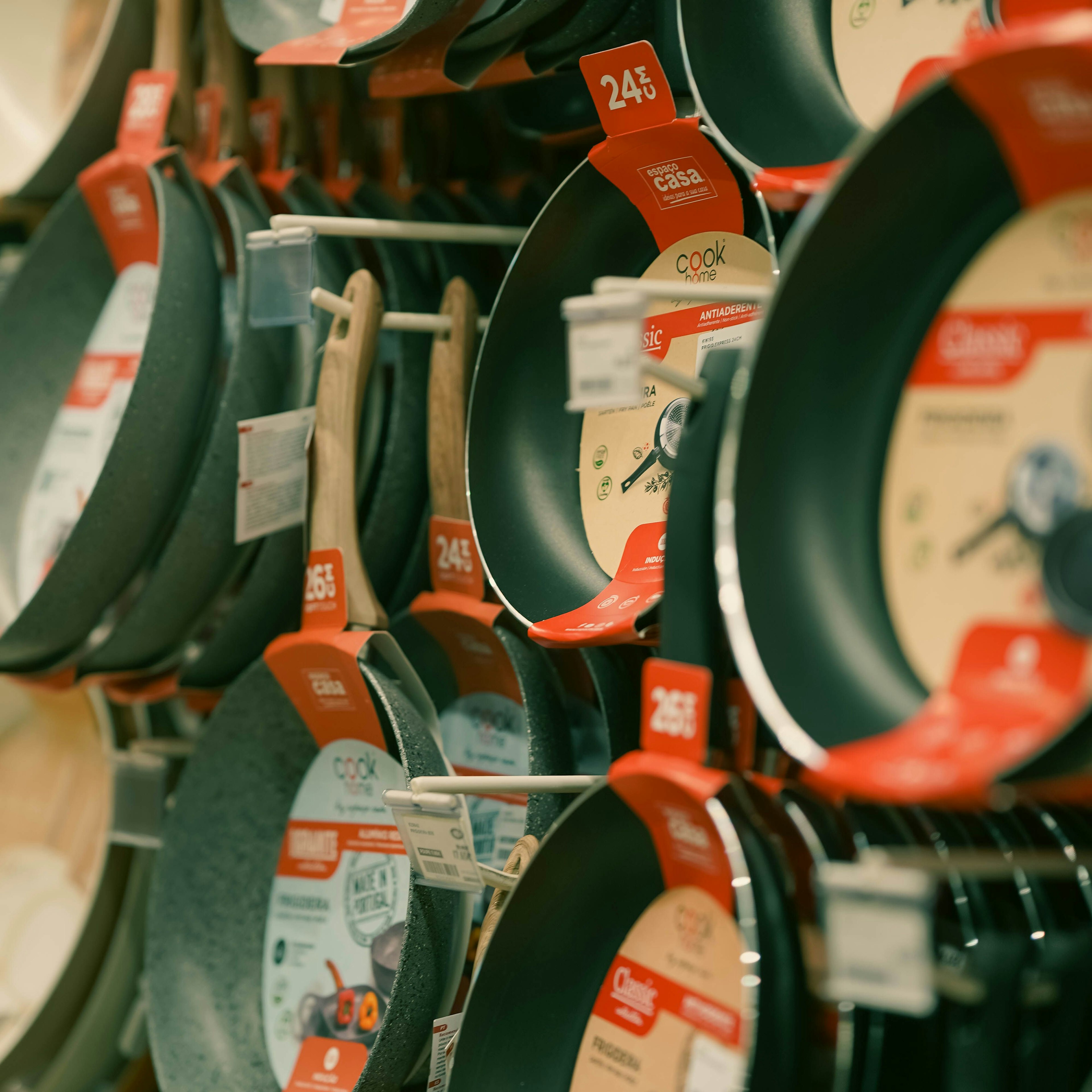 Dans un magasin d'ustensiles de cuisine, un présentoir mural où sont accrochées, sur des tringles, des poêles antiadhésives de différentes tailles, chacune munie d'une étiquette de prix rouge.