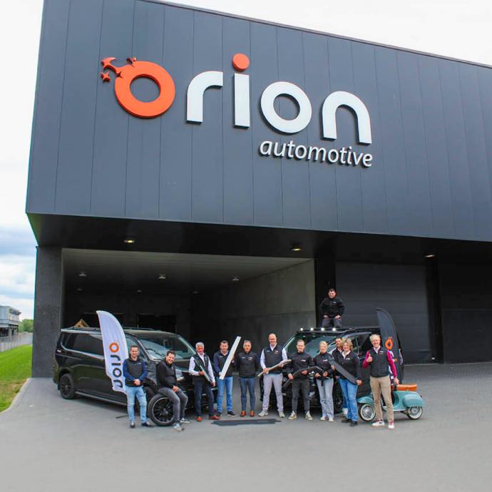 Une rangée de camionnettes de livraison blanches de bpost, aménagées par Orion Automotive, était garée sur un terrain en gravier sous un ciel couvert.