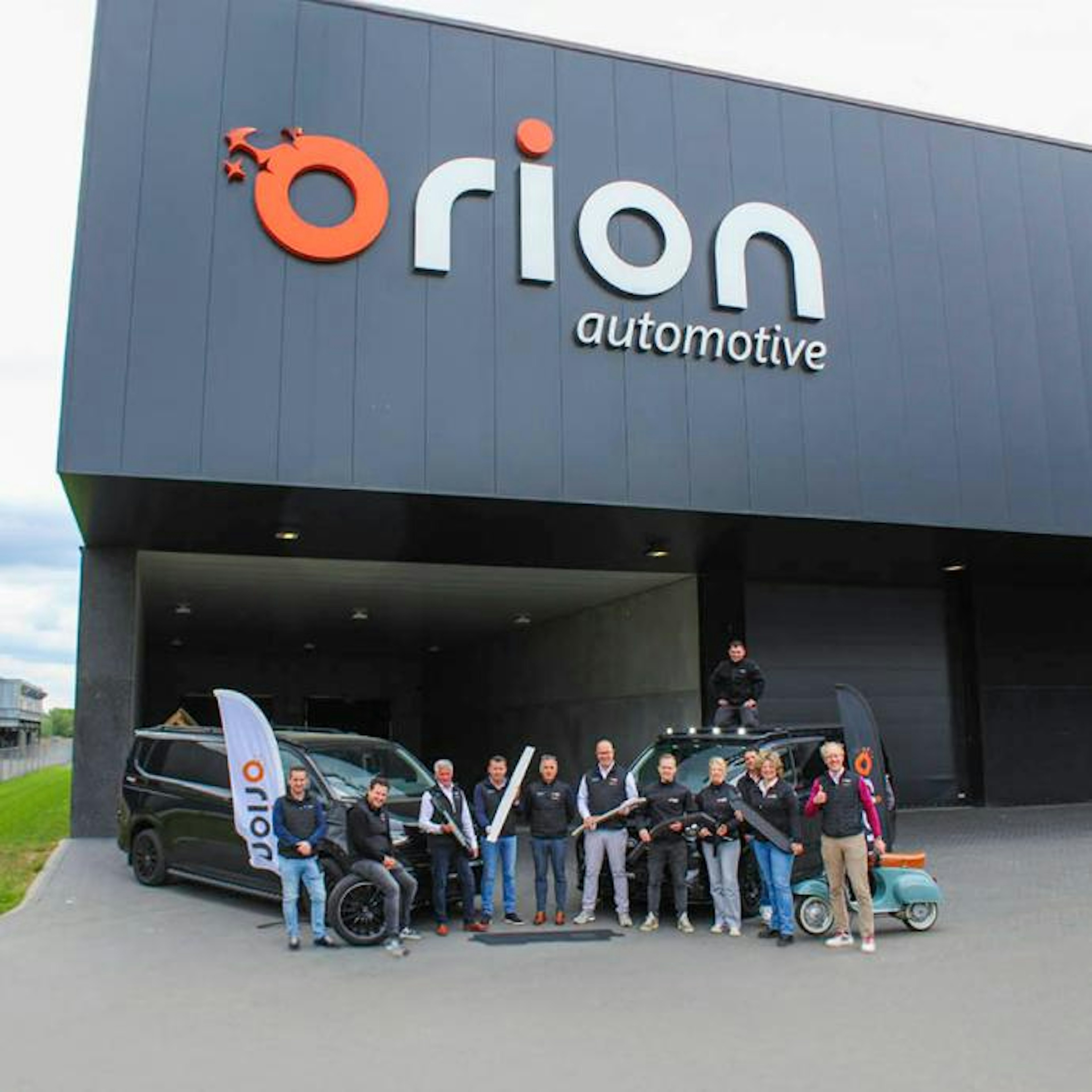 Une rangée de camionnettes de livraison blanches de bpost, aménagées par Orion Automotive, était garée sur un terrain en gravier sous un ciel couvert.