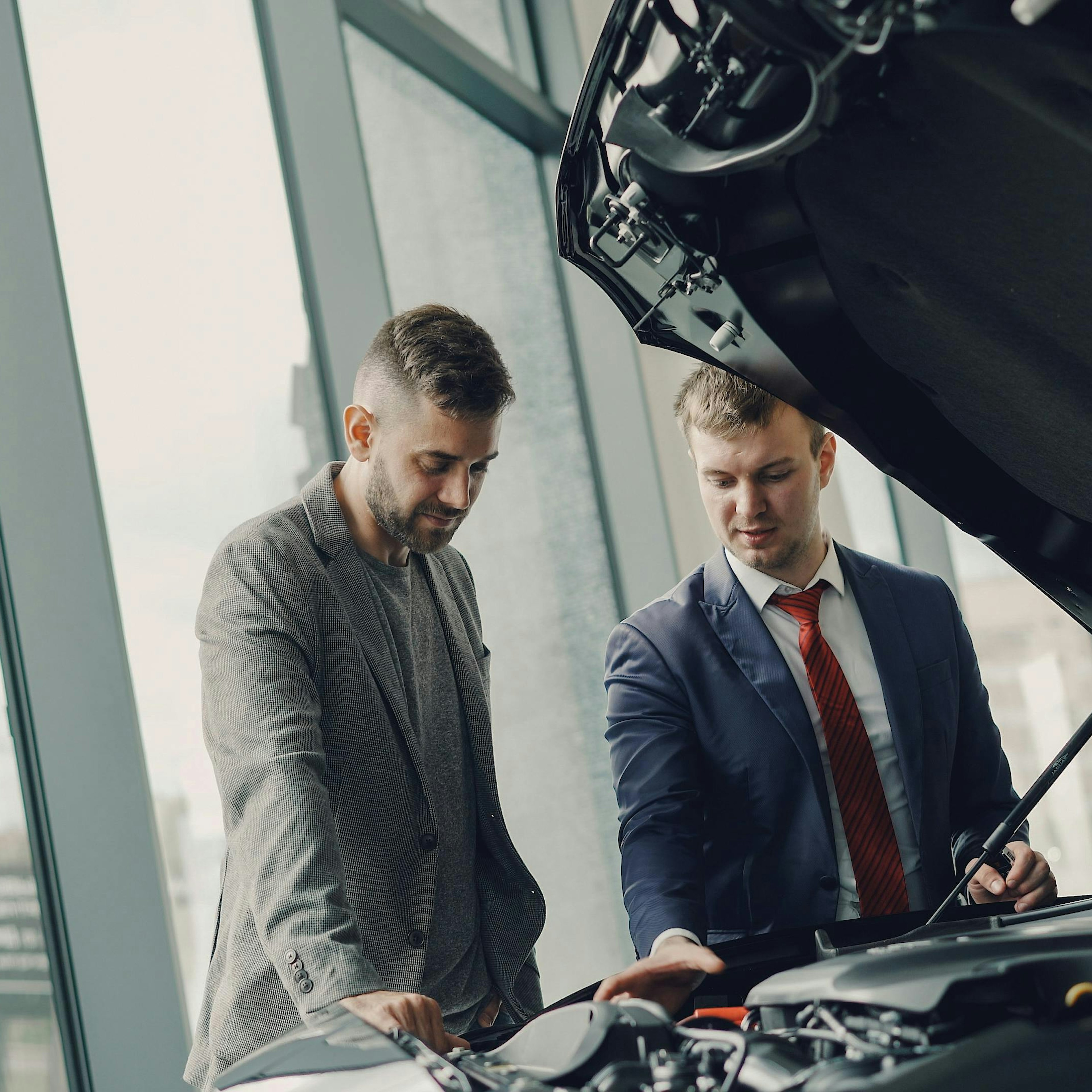 Dans le showroom vitré d'un concessionnaire, un vendeur en costume montre à un client le compartiment moteur d'une voiture dont le capot est relevé.