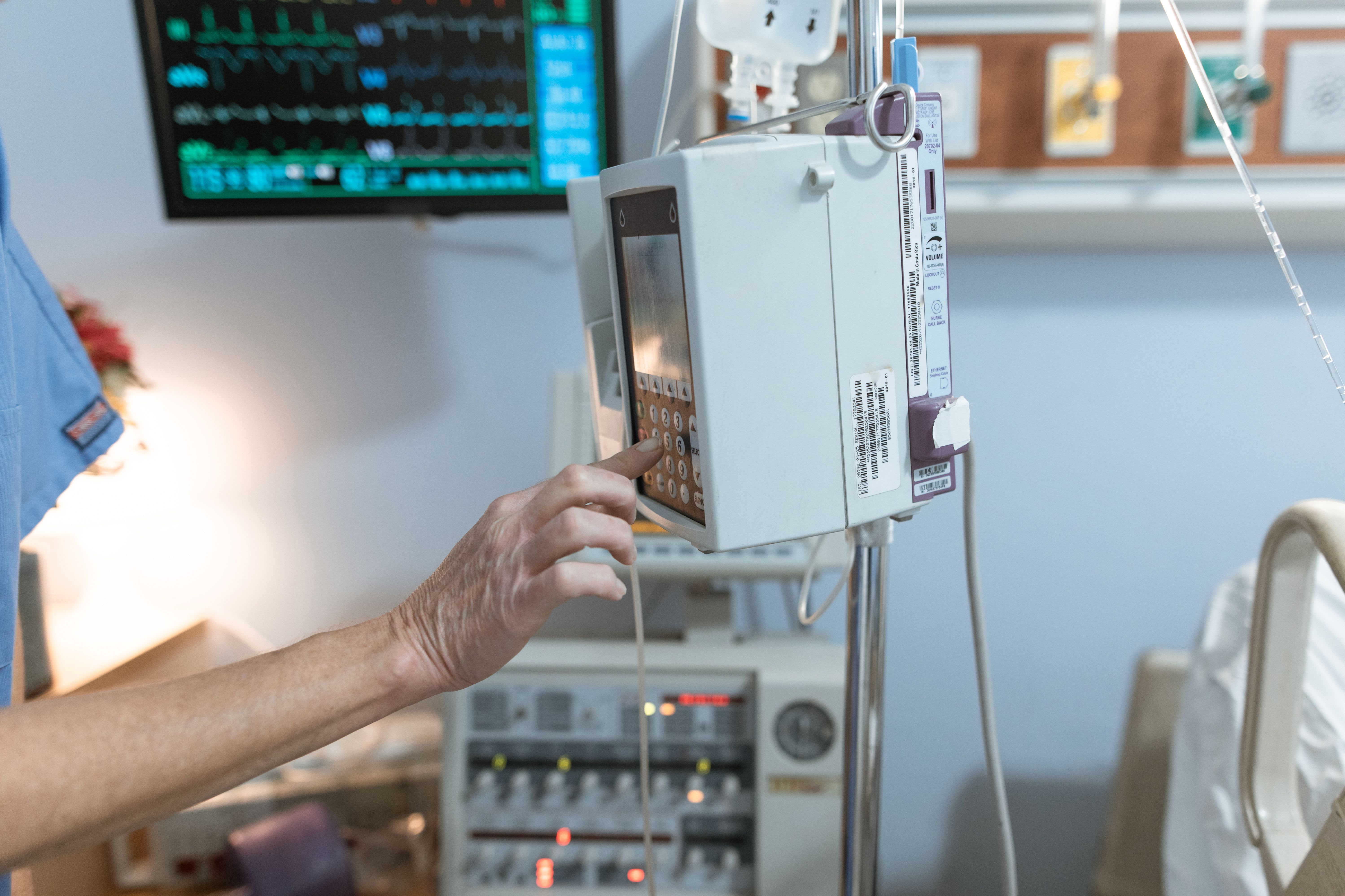 Une infirmière vêtue d'une blouse bleue règle le débit d'une pompe à perfusion intraveineuse à l'hôpital, tandis qu'un moniteur de surveillance affiche les courbes des signes vitaux en arrière-plan.