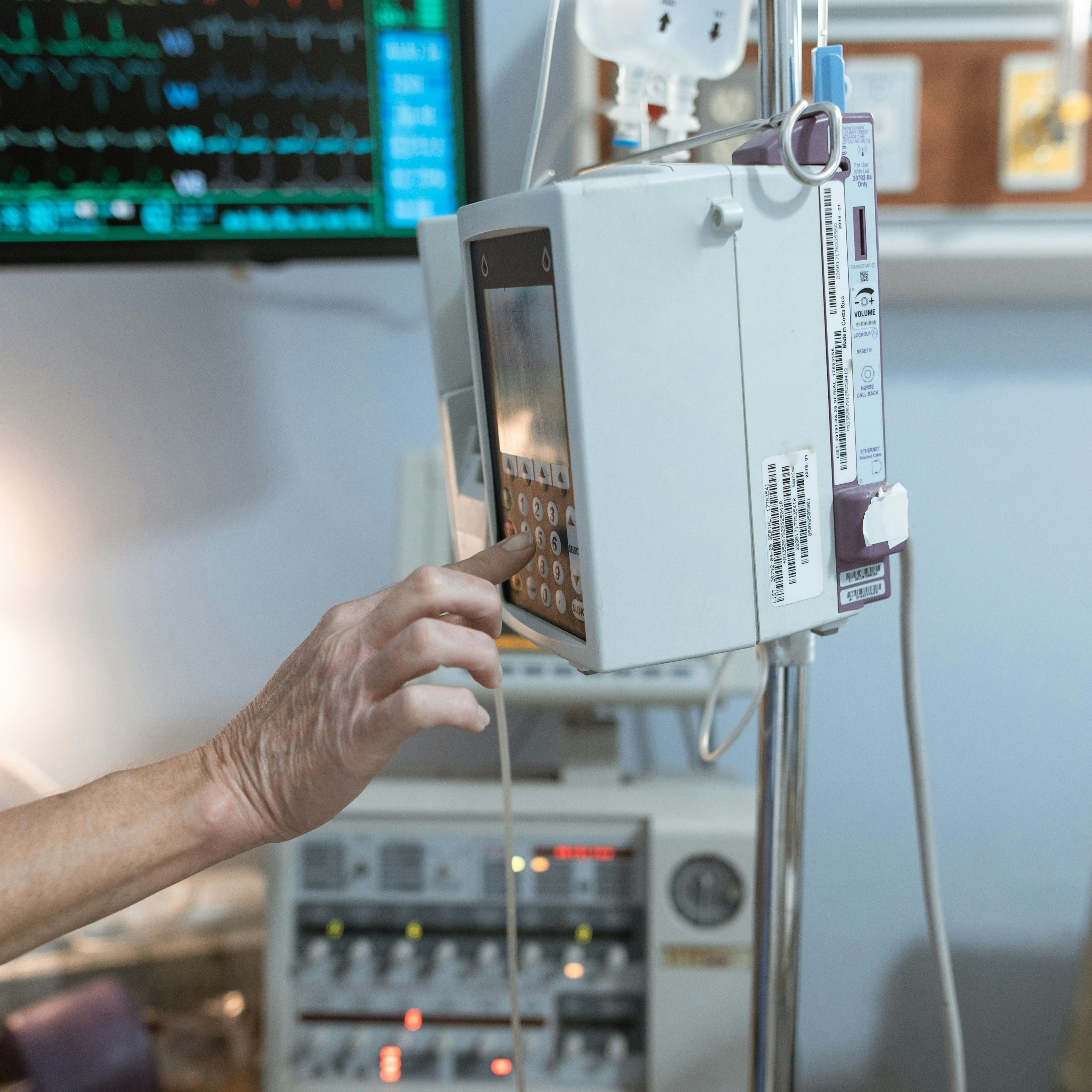 A nurse in blue scrubs sets the rate on a hospital IV infusion pump, with a patient monitor showing vital-sign traces in the background.
