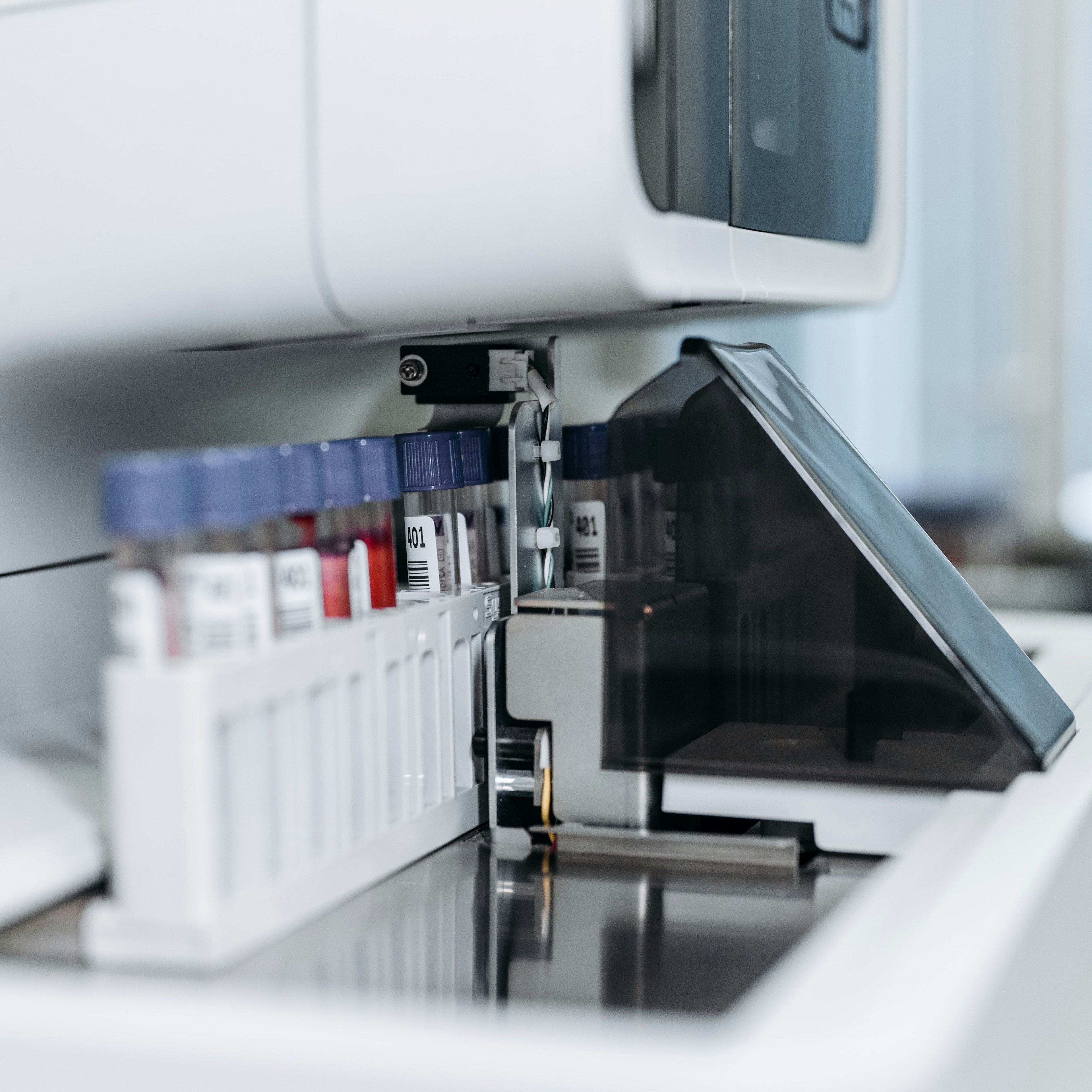 Close-up of an automated lab analyser with a loading tray of barcoded sample tubes, one capped in blue and one filled with red liquid.