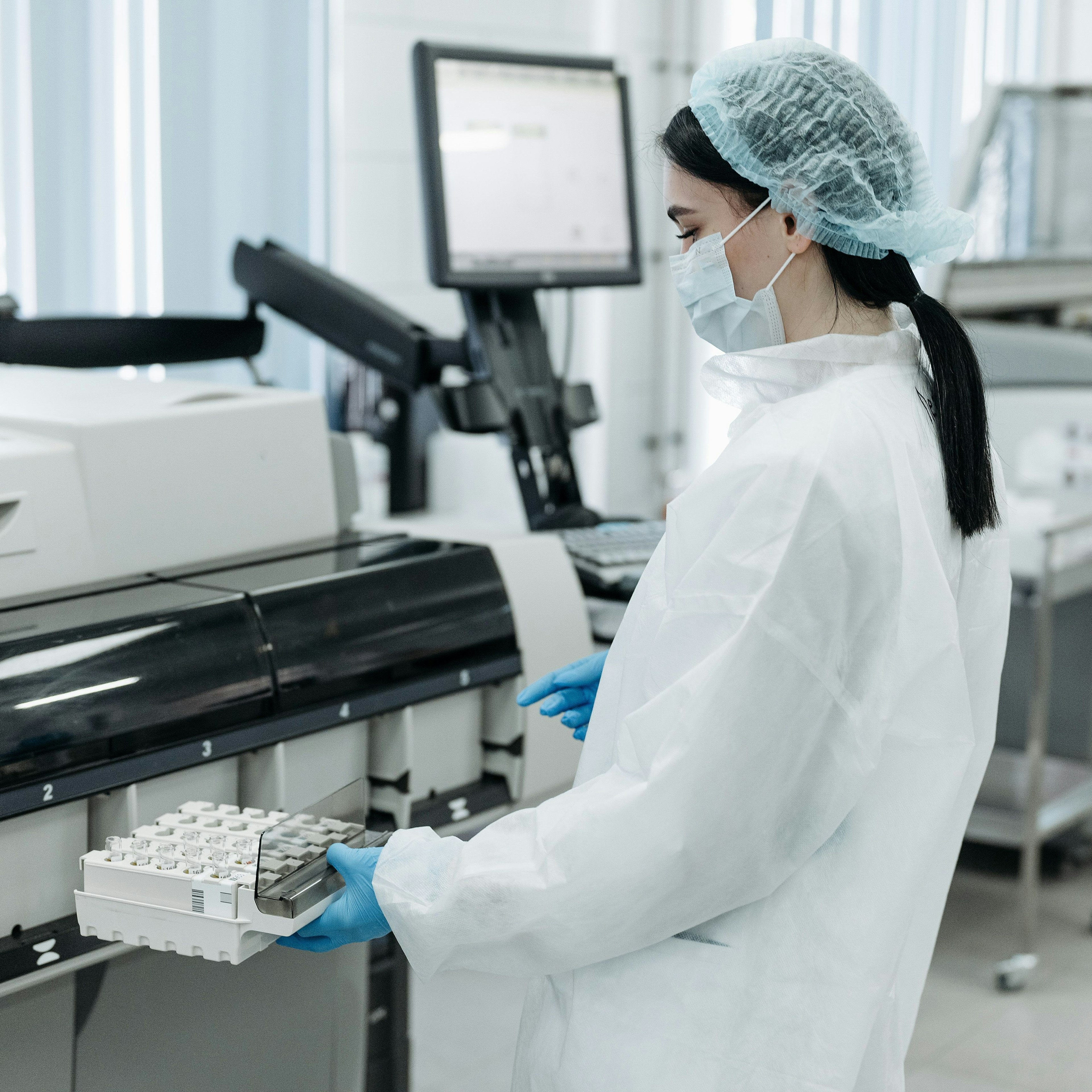 Une technicienne de laboratoire travaillant dans un laboratoire clinique moderne, vue de profil. Elle porte une blouse jetable blanche, un filet à cheveux, un masque chirurgical et des gants en nitrile bleus ; ses longs cheveux foncés sont attachés en queue de cheval. Elle tient un portoir à échantillons blanc rempli de tubes, qu'elle charge dans un grand analyseur automatisé doté de tiroirs numérotés (de 1 à 6). Derrière elle se trouvent des écrans montés sur des bras articulés et d'autres équipements de laboratoire, le tout dans un environnement lumineux, propre, aux tons blancs et froids.