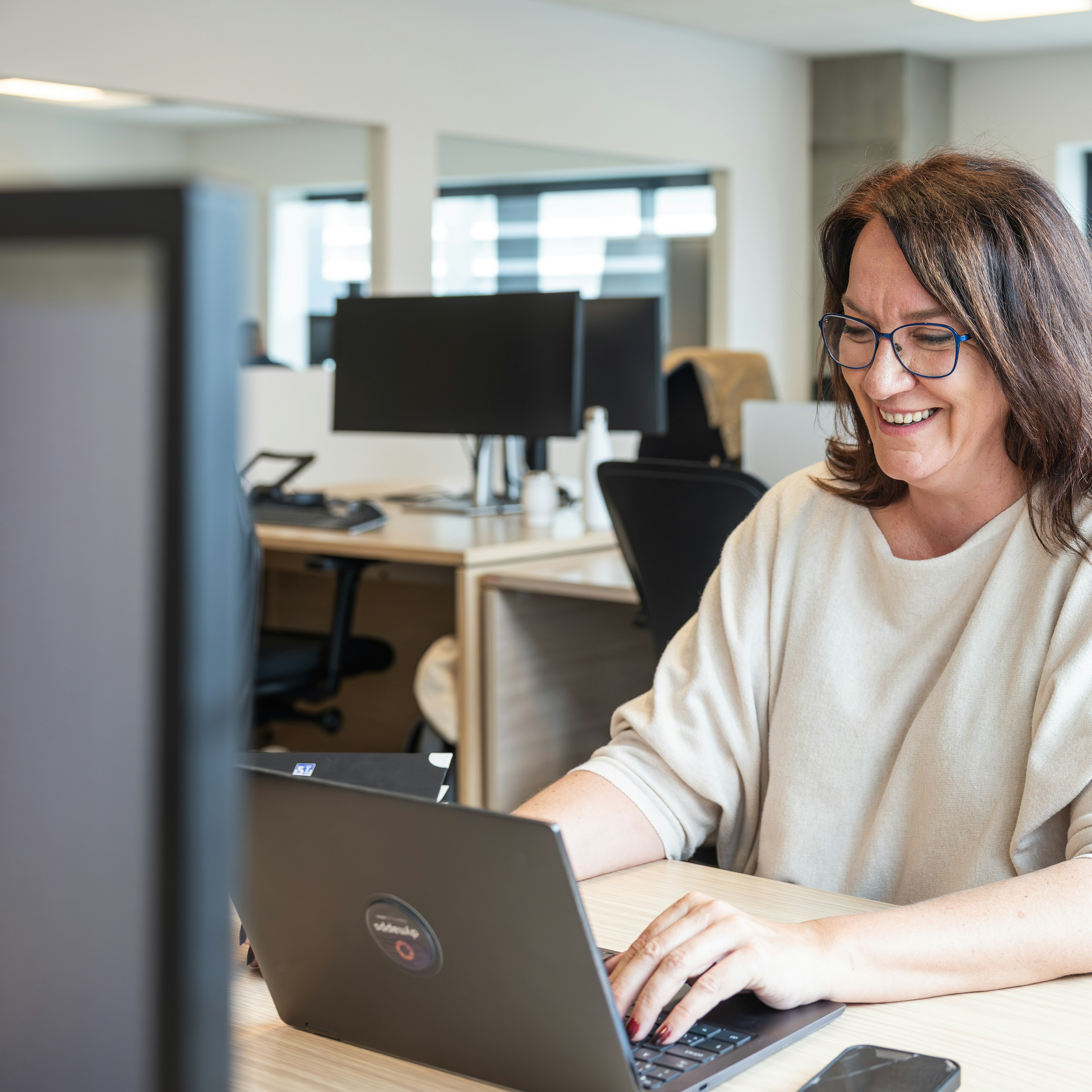Une femme souriante portant des lunettes, en train de taper sur un ordinateur portable