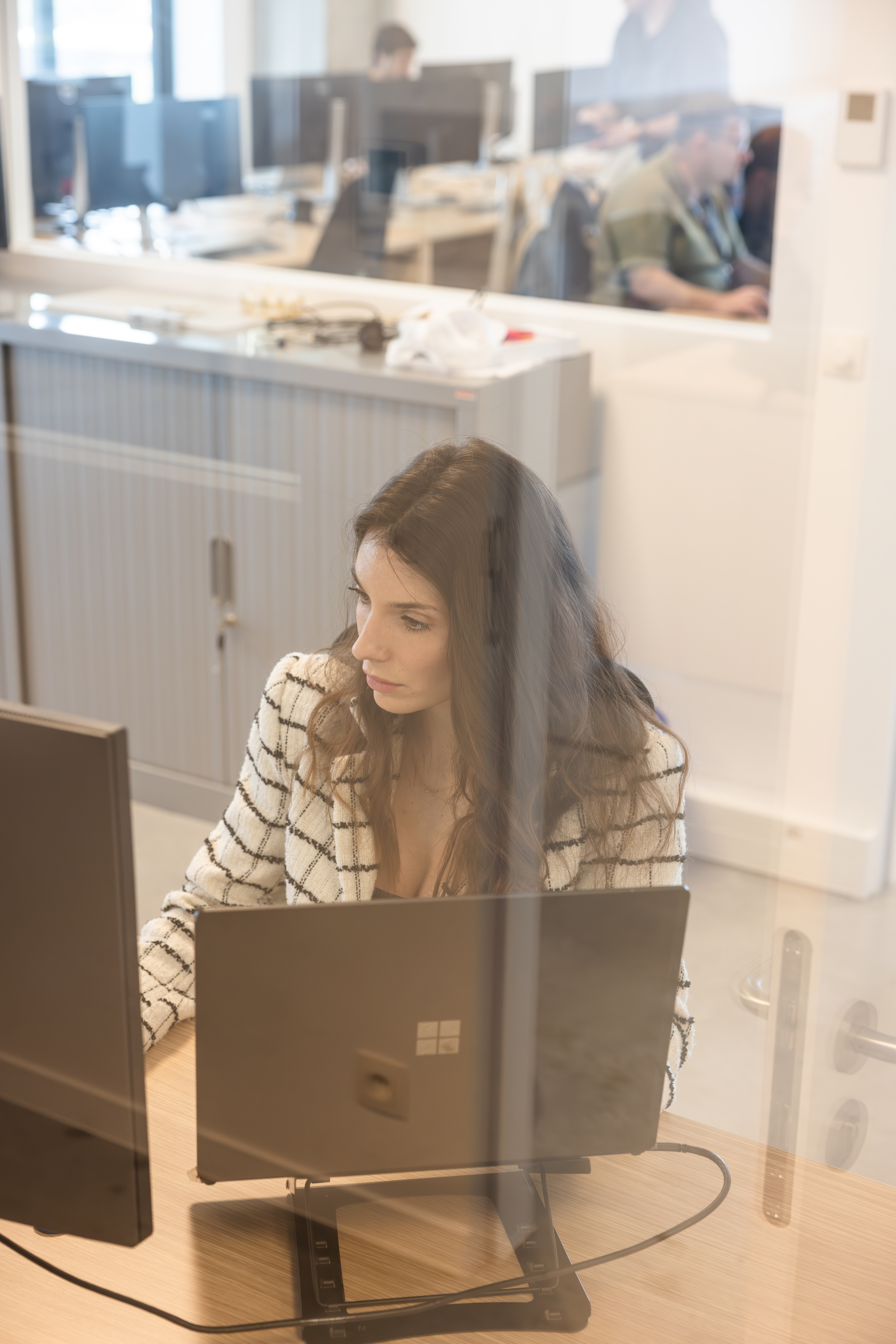 Une femme concentrée sur son ordinateur portable, vue à travers la vitre d'un bureau