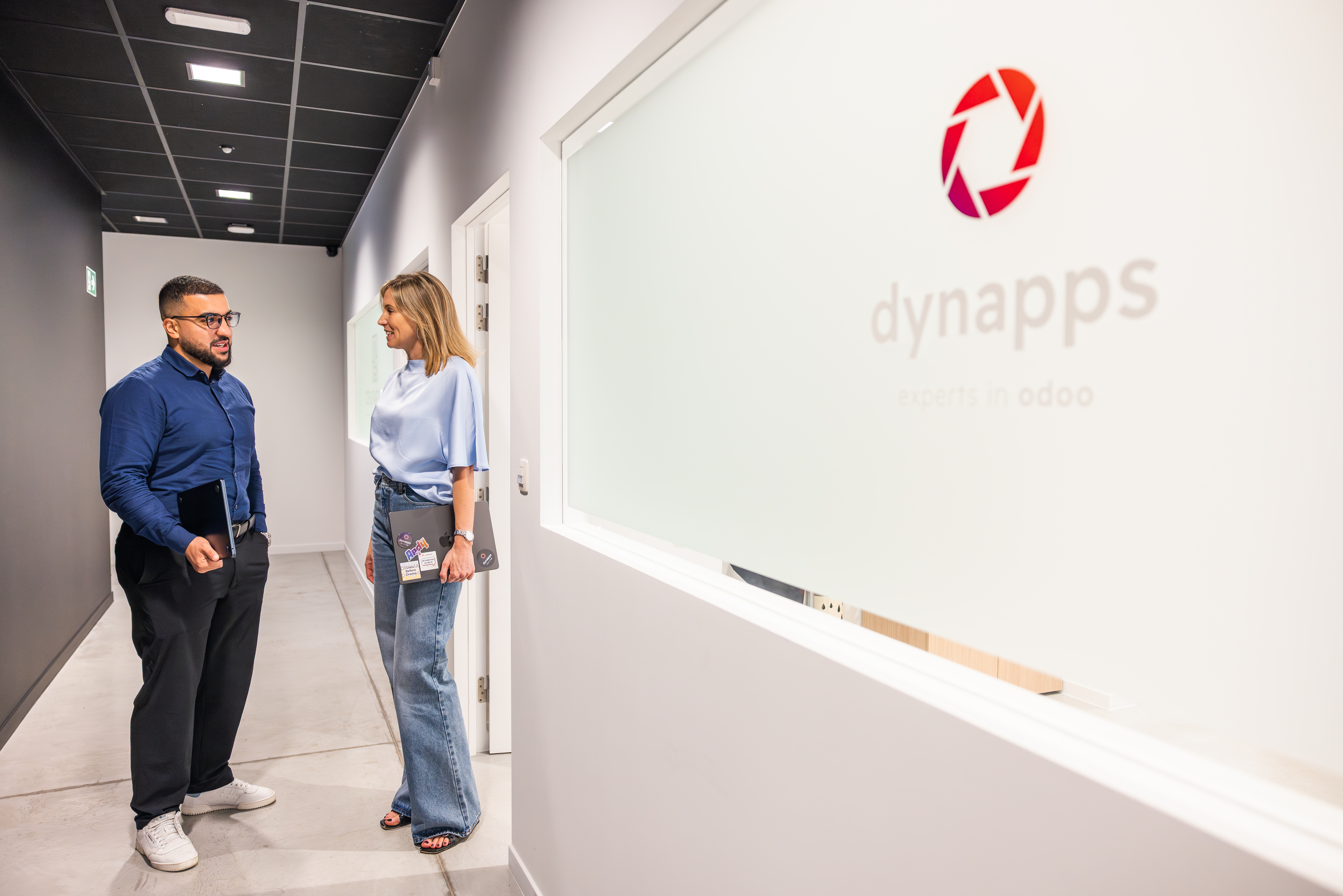 Two professionals talking in a modern office hallway, with the Dynapps logo and name visible on the wall.