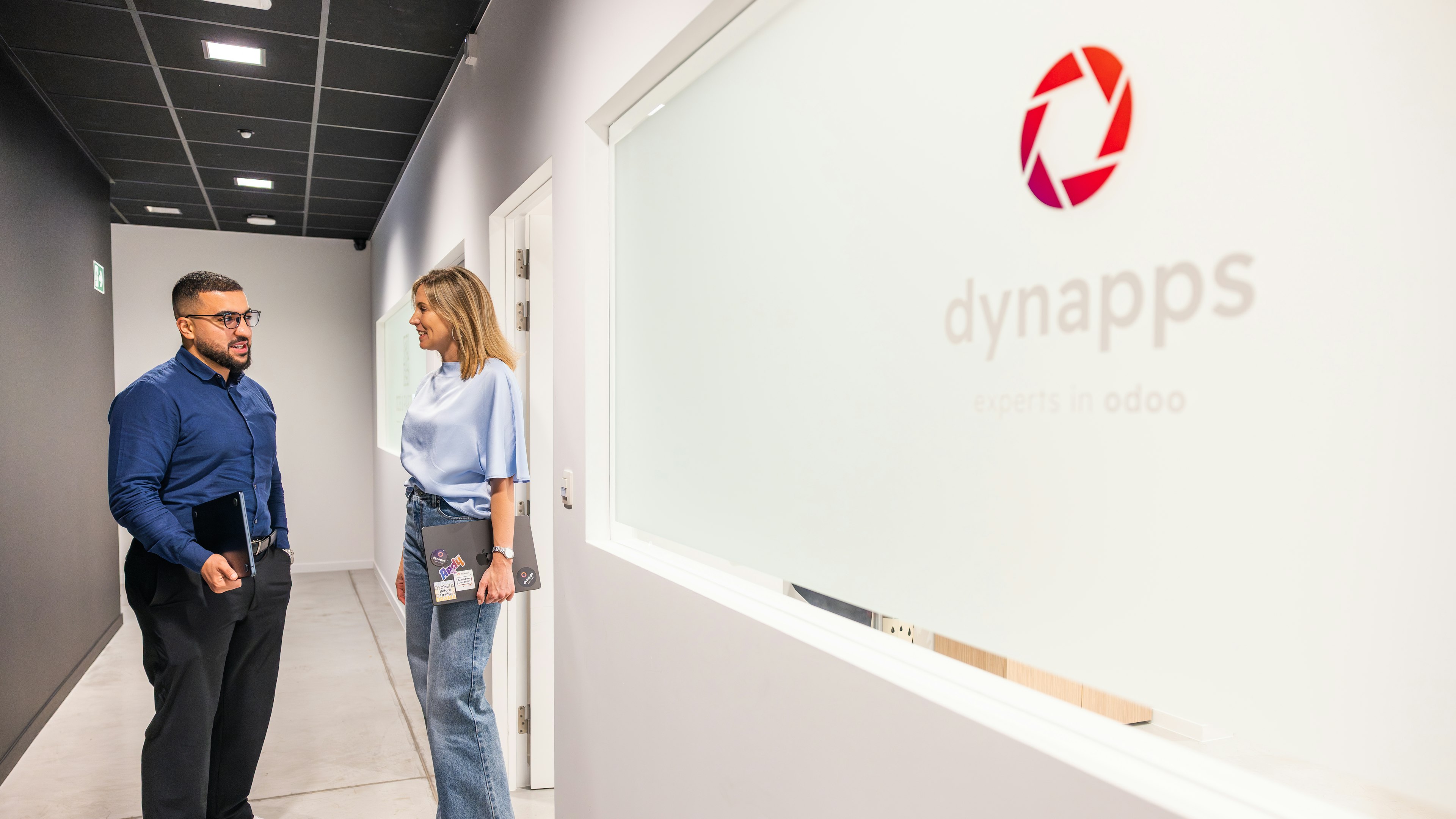Two professionals talking in a modern office hallway, with the Dynapps logo and name visible on the wall.