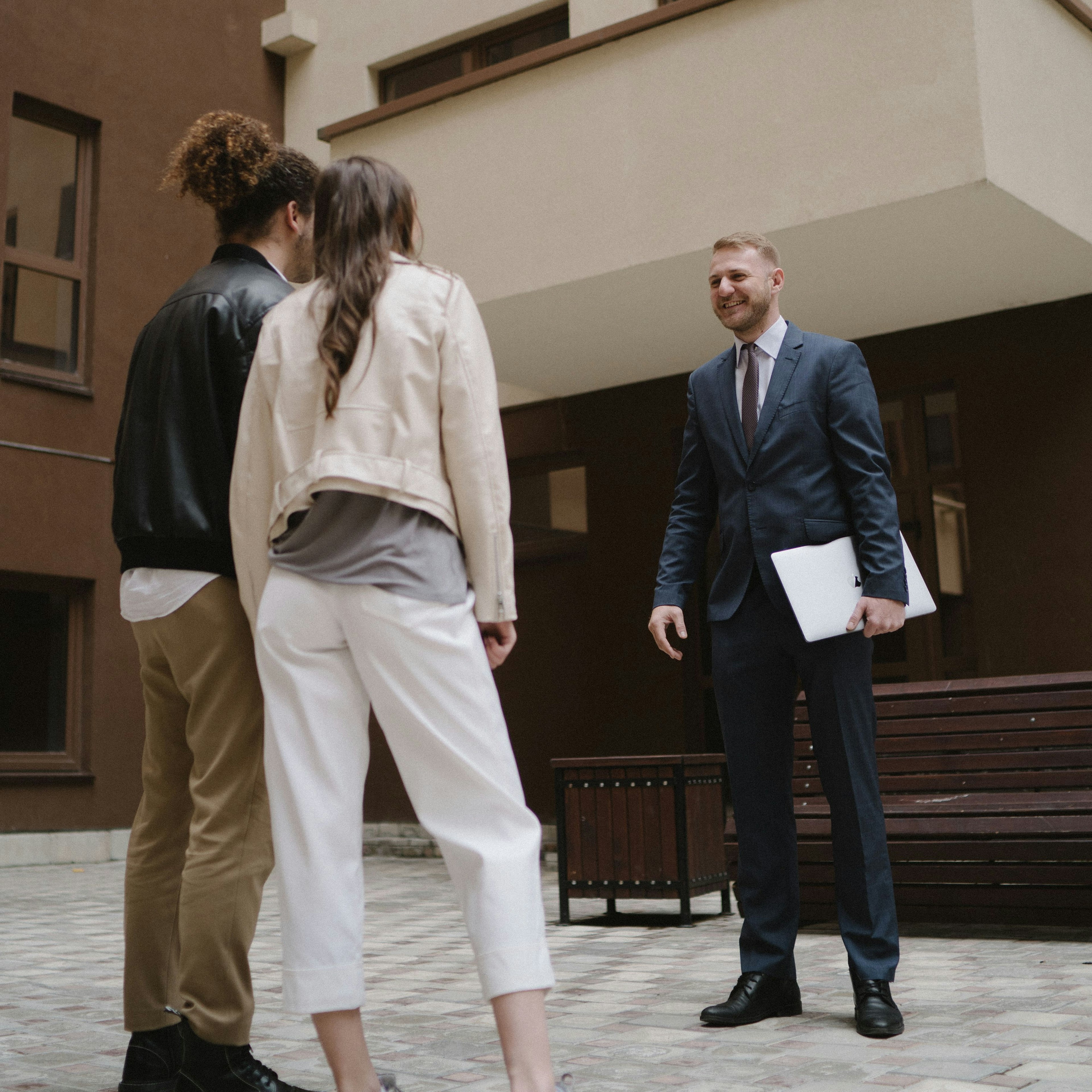 A smiling agent in a suit holds a laptop while greeting a young couple in the paved courtyard of a residential building.