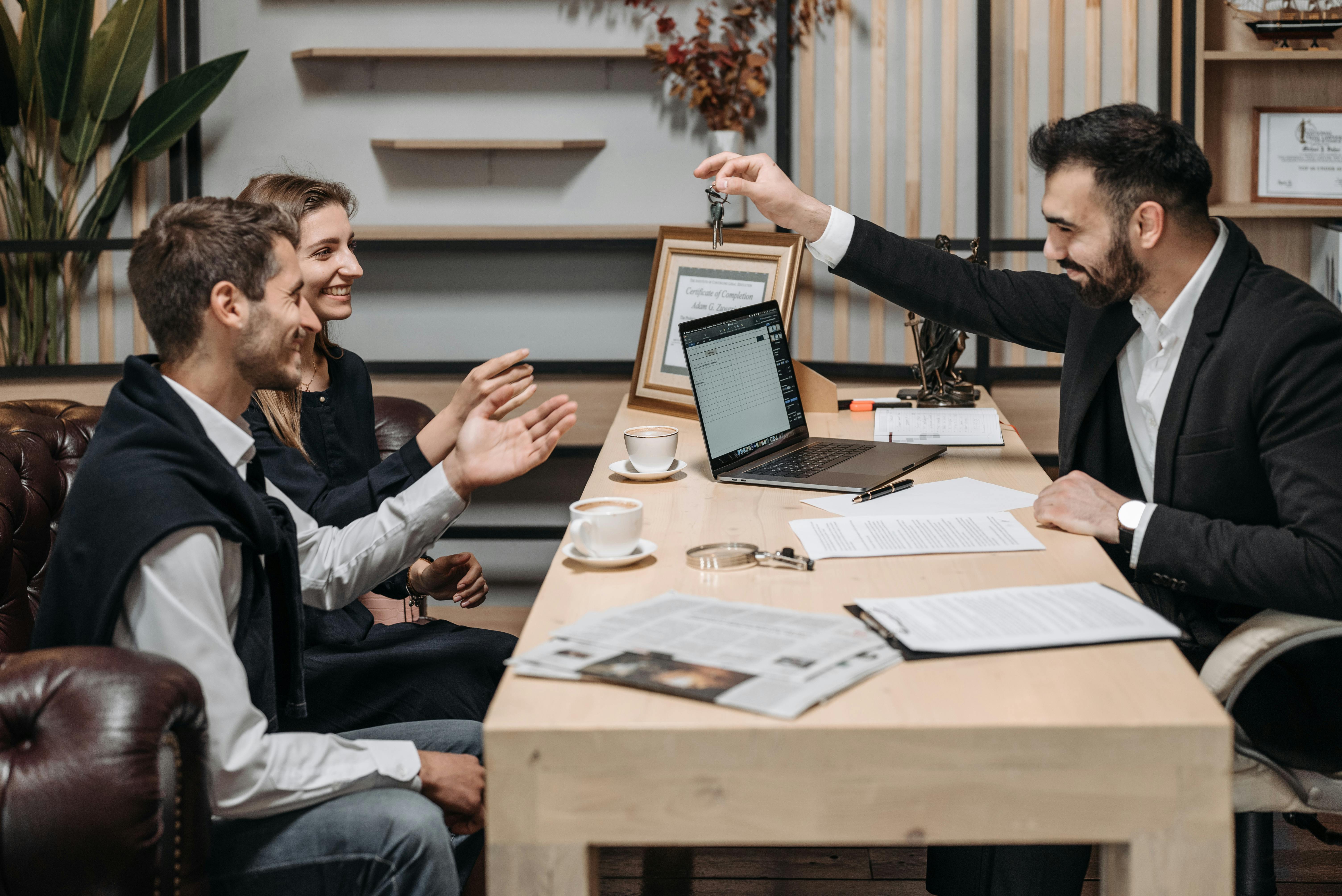 A smiling agent hands a set of keys across his desk to a young couple during a meeting, with a laptop, contract papers and coffee cups on the table.