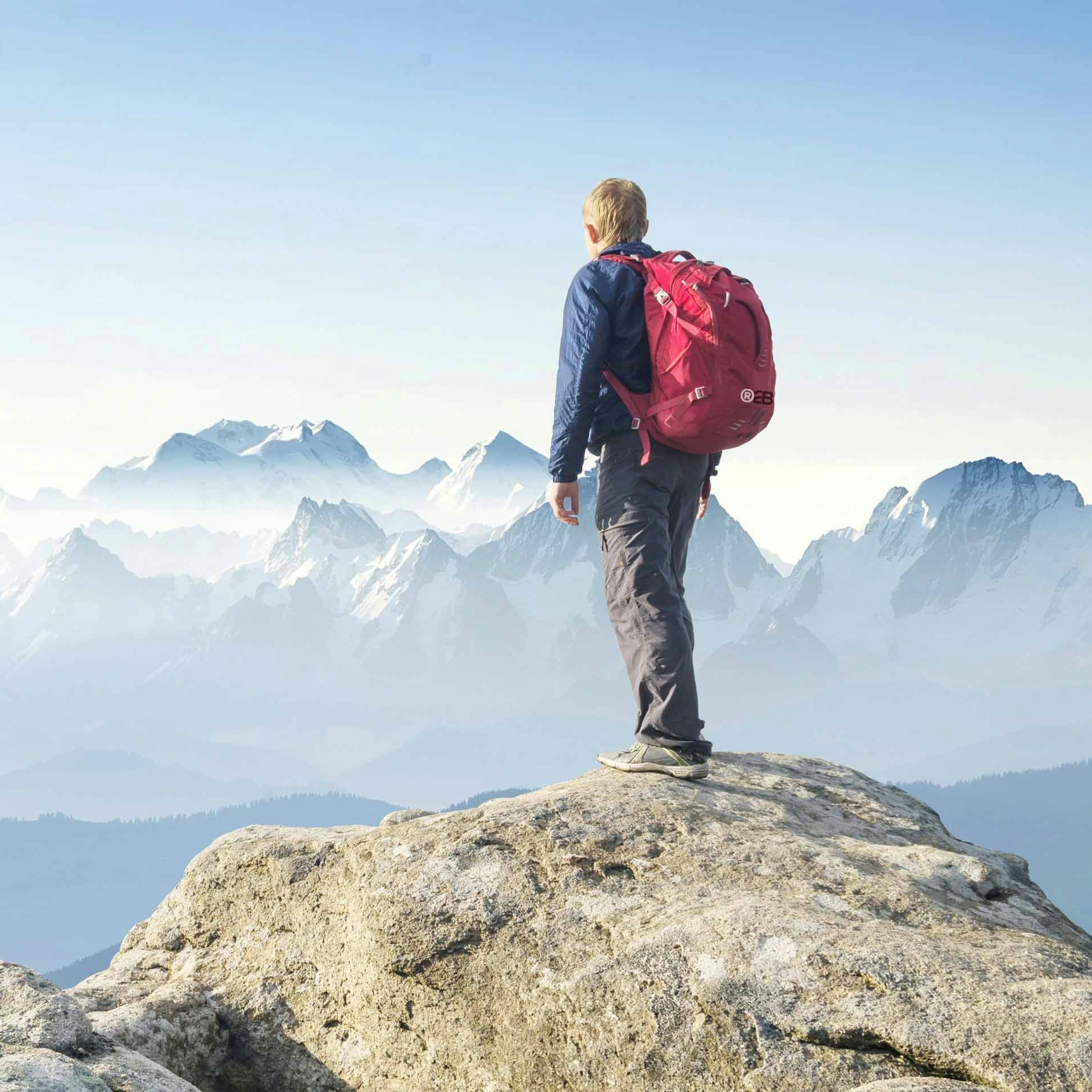 A hiker with a red backpack stands on a rocky summit, looking out over a range of misty, snow-capped mountains.