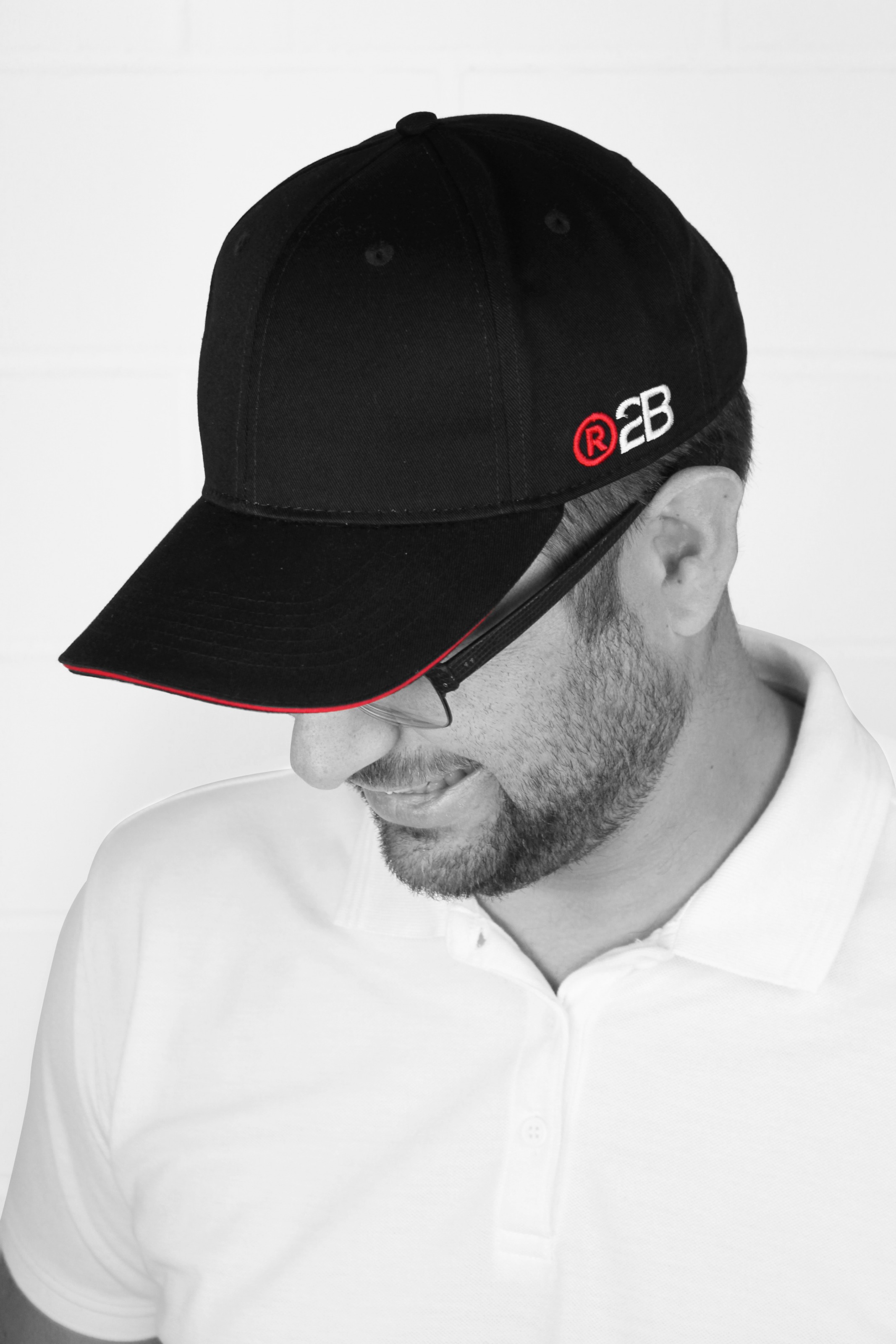 A man in a white polo shirt wears a black branded baseball cap, photographed from the side in black and white against a white wall.