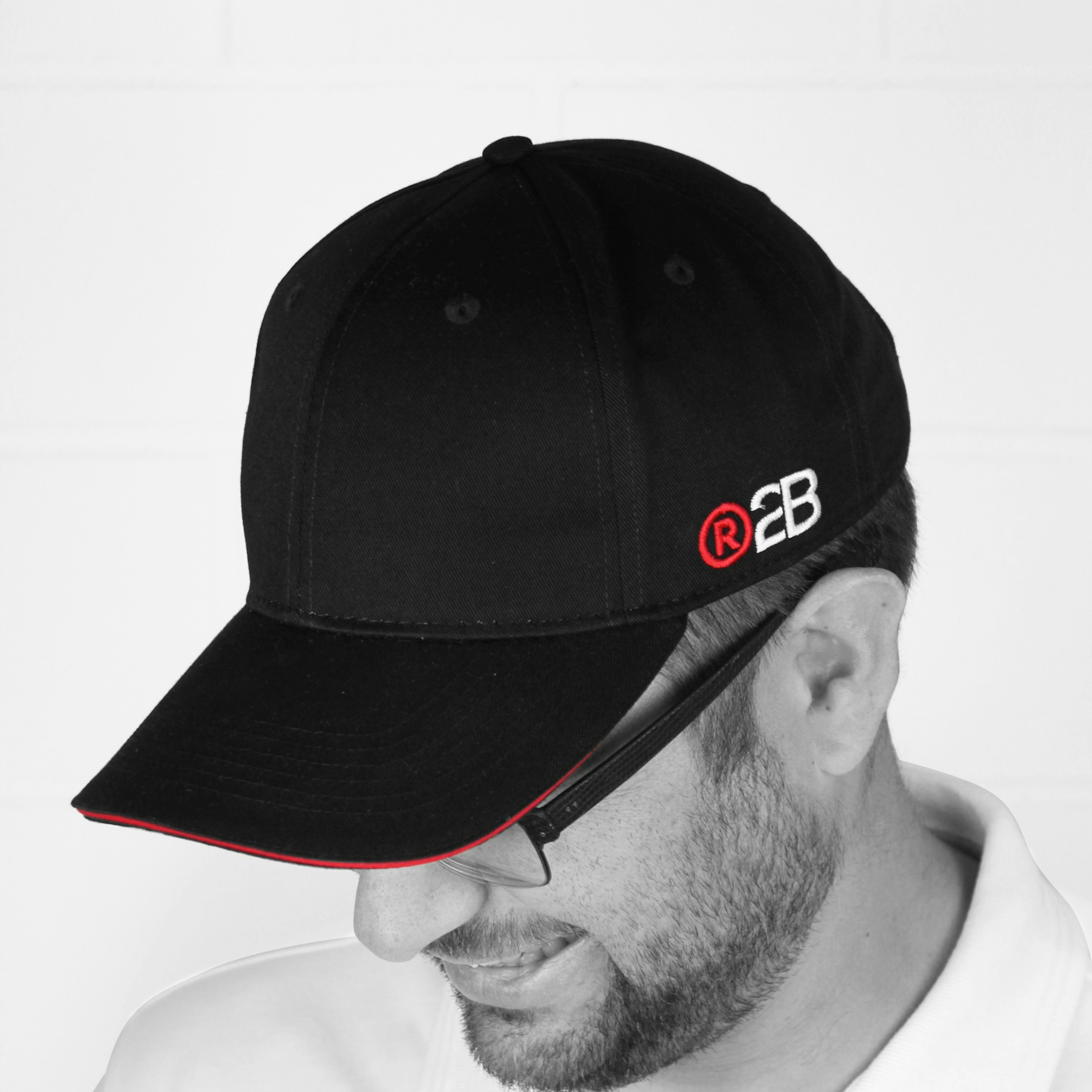 A man in a white polo shirt wears a black branded baseball cap, photographed from the side in black and white against a white wall.