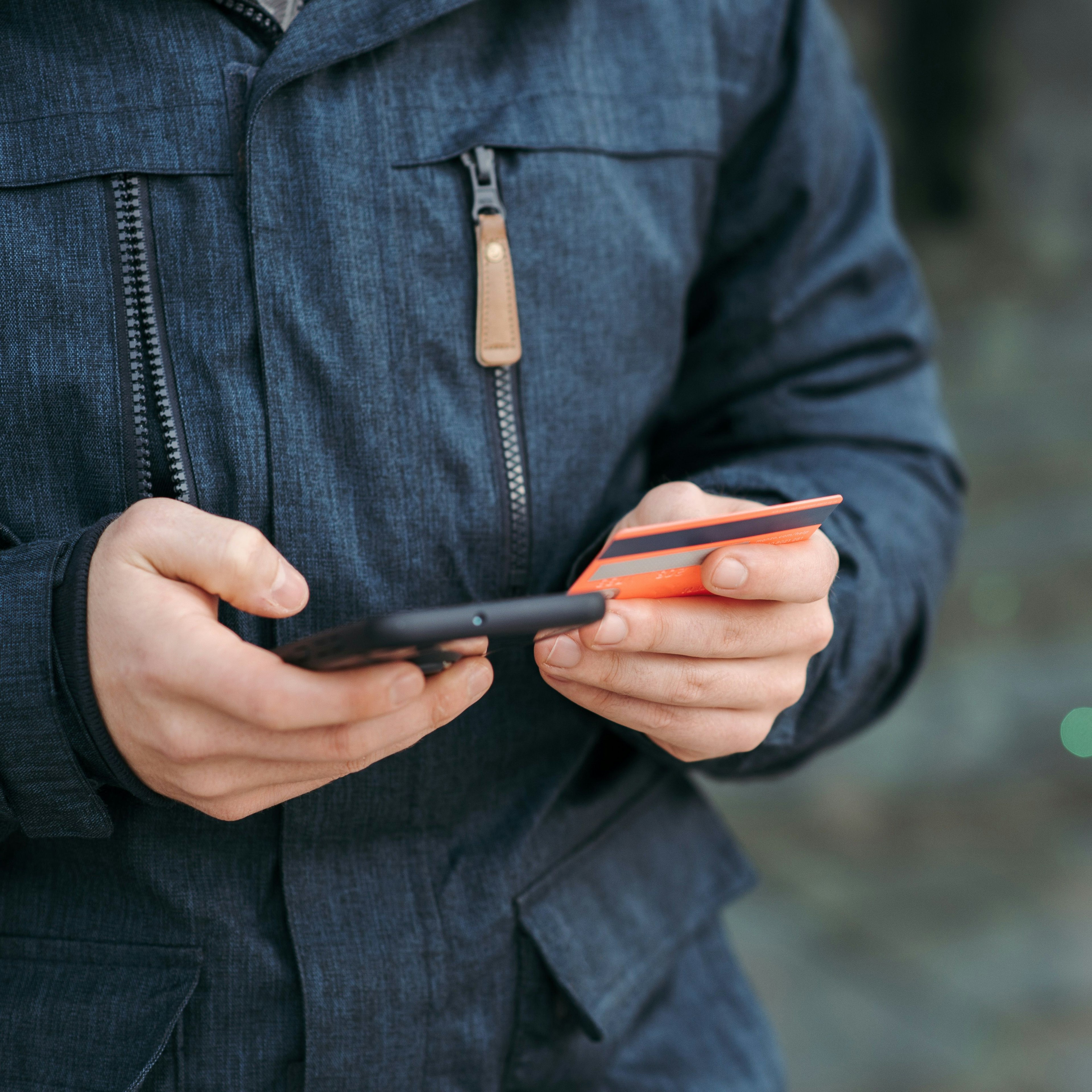 Una persona sostiene un teléfono inteligente y una tarjeta de crédito naranja al aire libre, realizando una transacción o pago digital móvil.