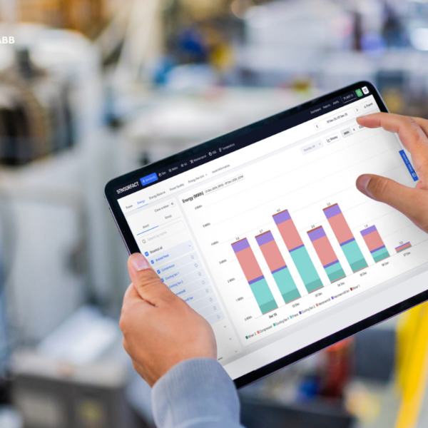 Gros plan sur des mains tenant une tablette affichant le tableau de bord de suivi énergétique Sensorfact, avec un histogramme, devant des équipements d'usine flous.