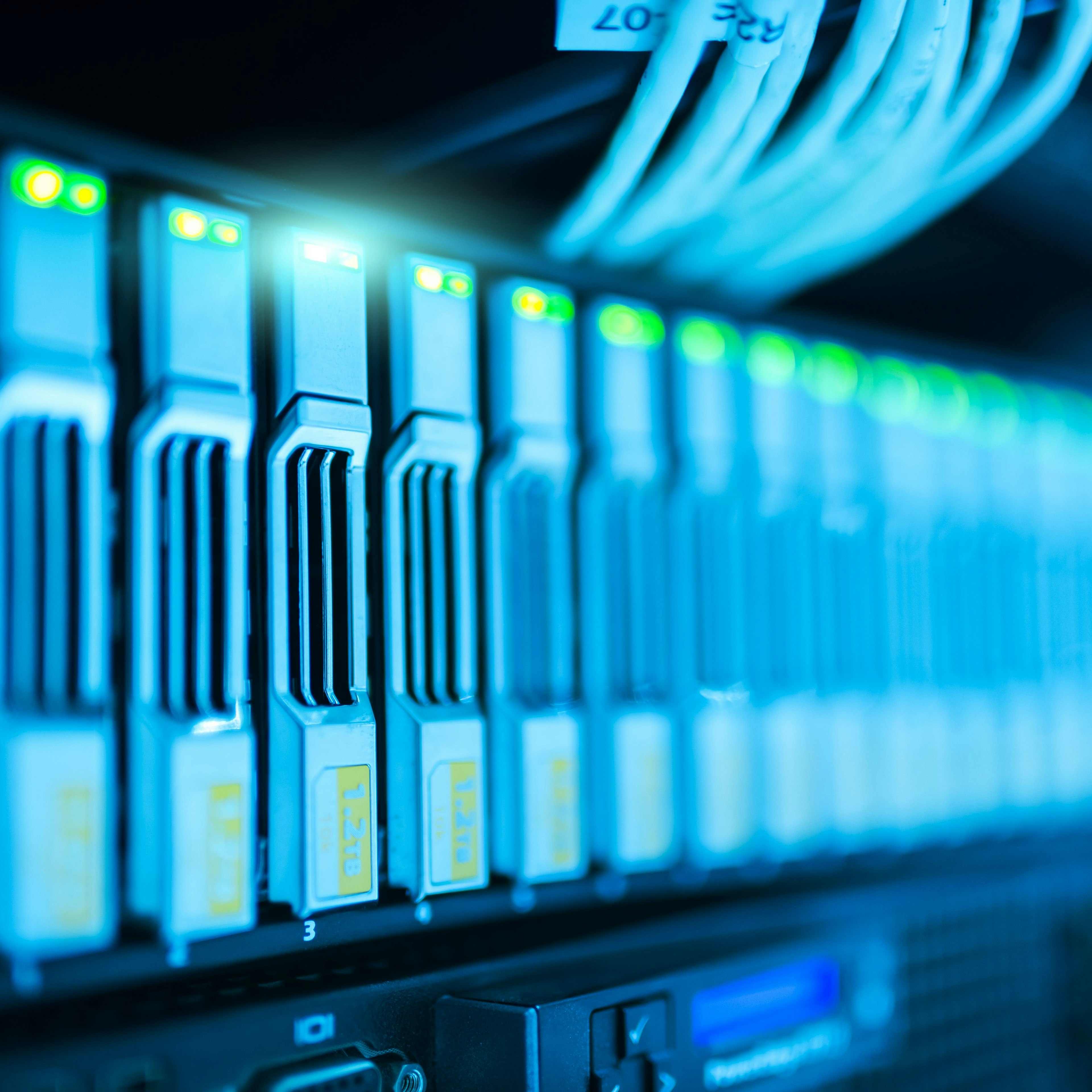 This image shows a close-up view of a data center server rack filled with multiple hard drive bays. The units feature glowing green status lights and connected network cables, all illuminated by a cool blue light.