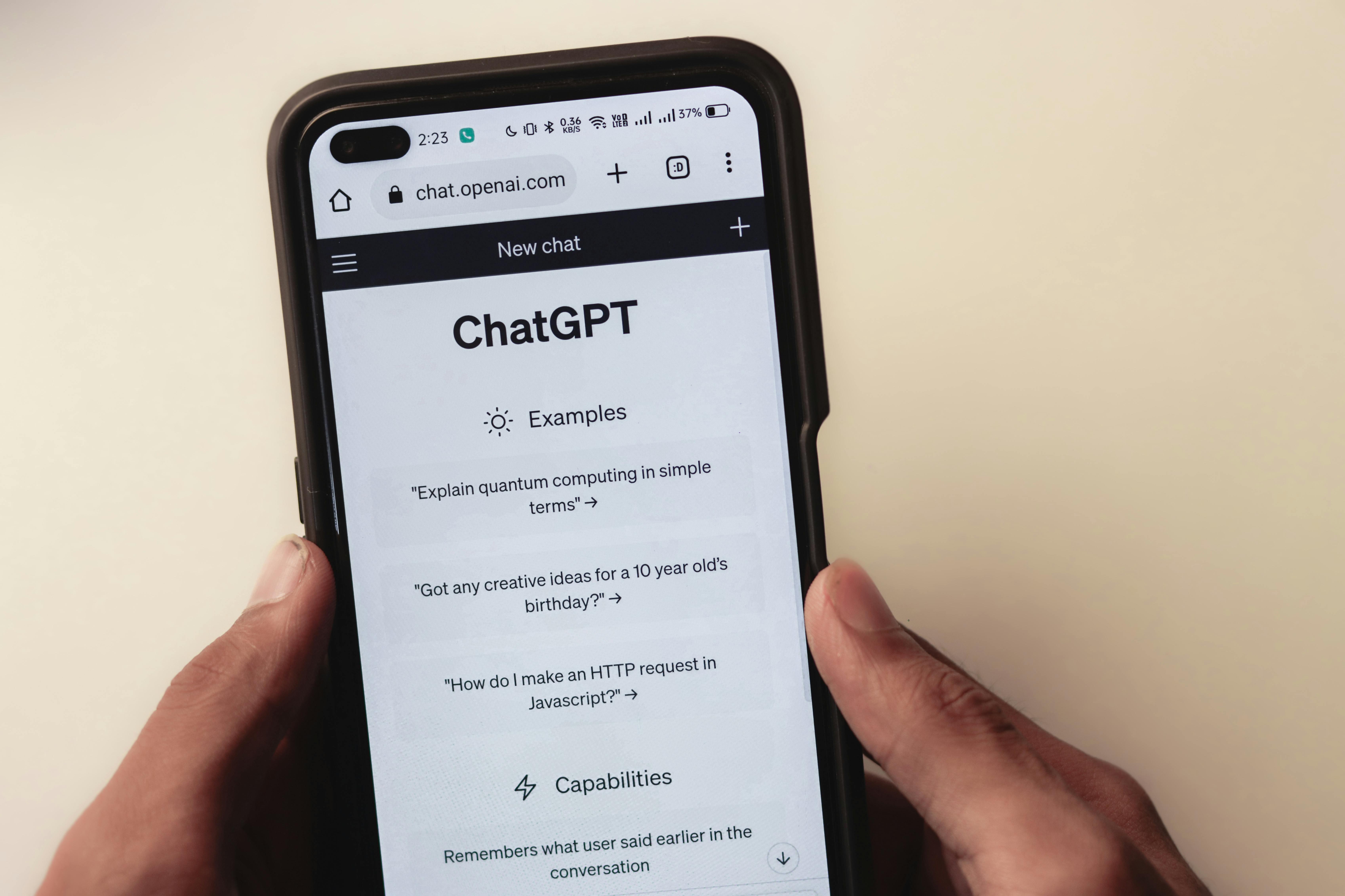 This image shows a person holding a smartphone displaying the ChatGPT mobile web interface, which includes sample prompts such as "Explain quantum computing in simple terms."