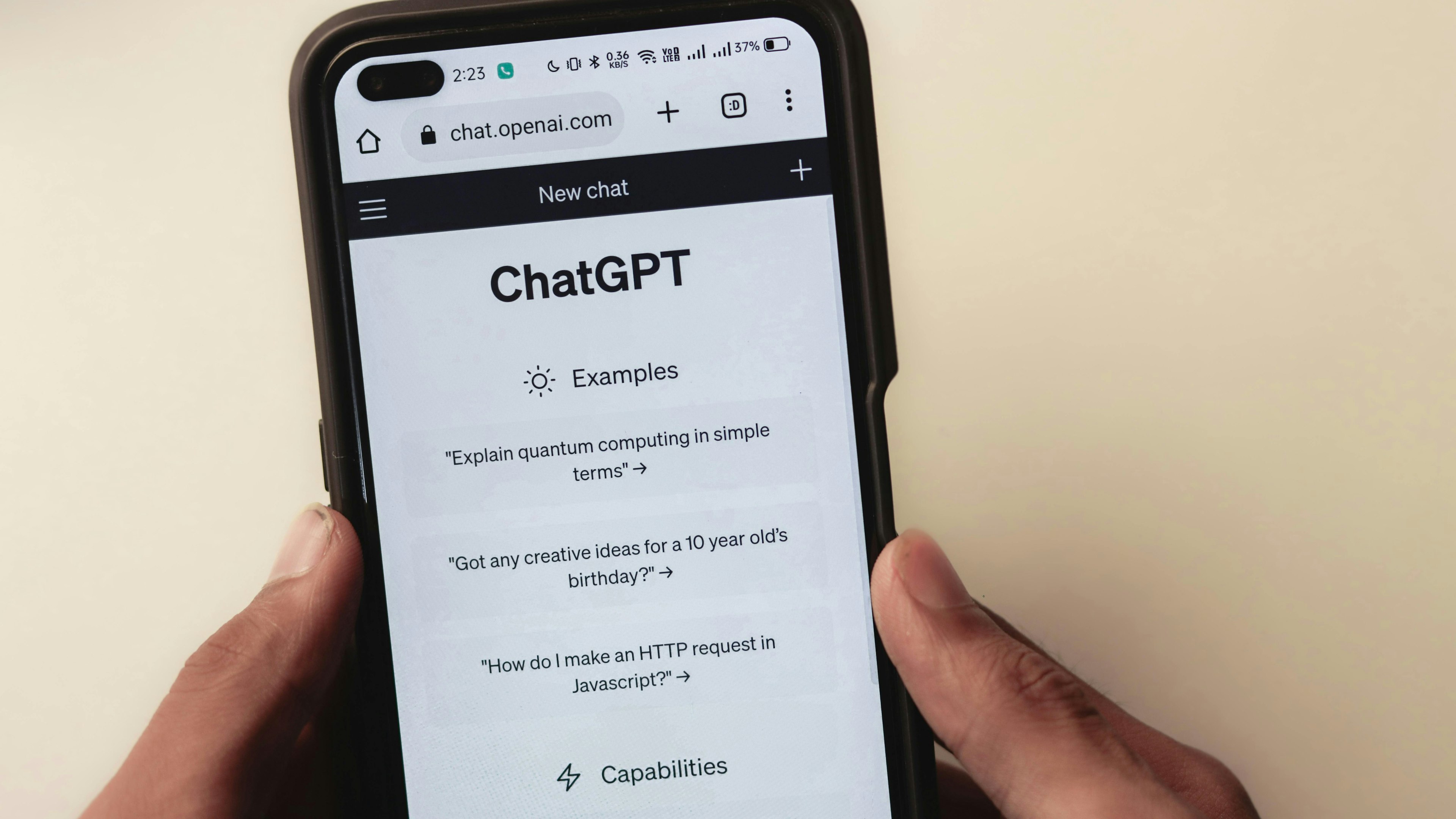 This image shows a person holding a smartphone displaying the ChatGPT mobile web interface, which includes sample prompts such as "Explain quantum computing in simple terms."
