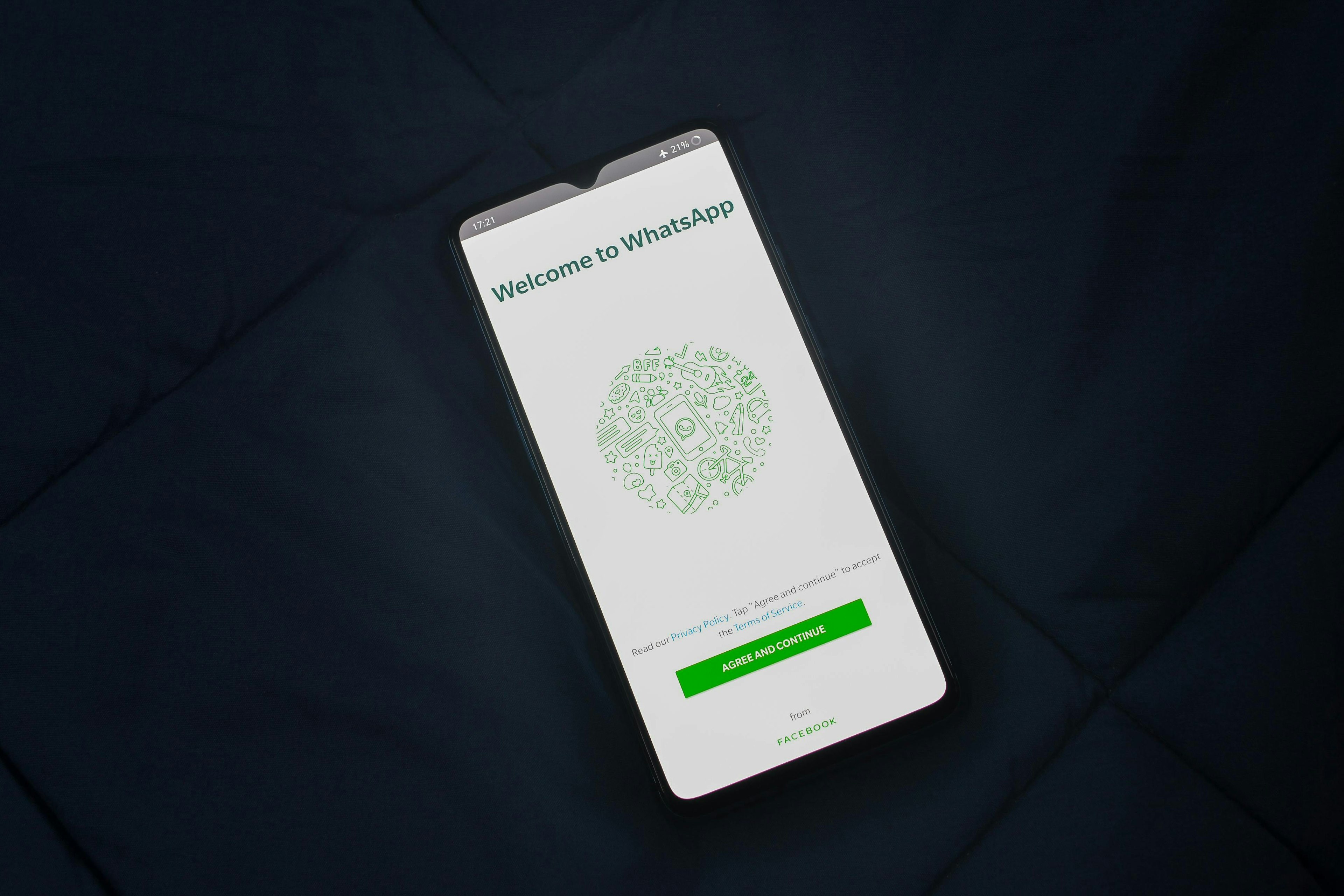This image shows a smartphone resting on a dark fabric surface, displaying the initial setup screen for the WhatsApp application with the text "Welcome to WhatsApp" and an "AGREE AND CONTINUE" button.