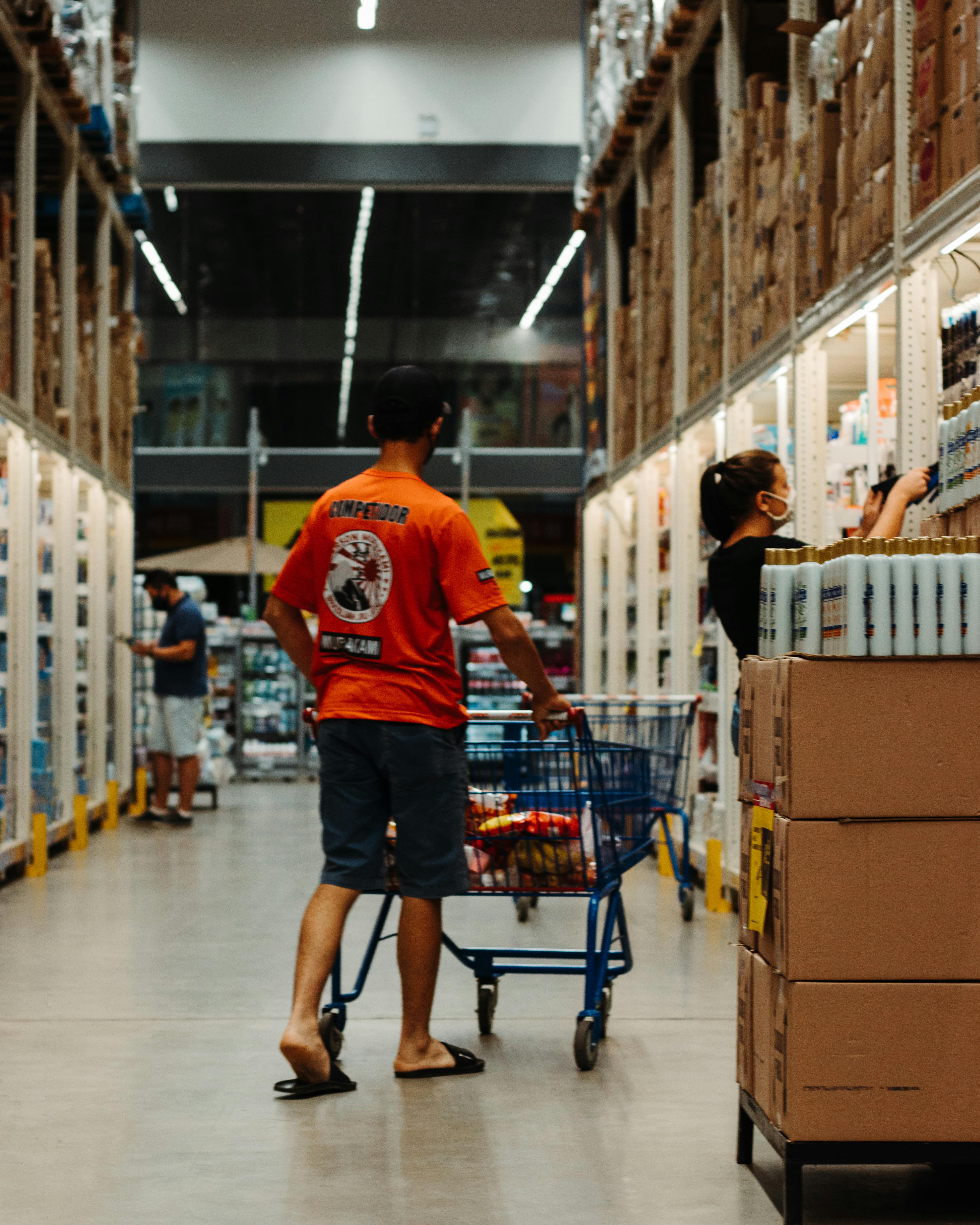 Un client pousse un chariot dans l'allée d'un magasin de type entrepôt, où les rayons de chaque côté sont chargés de marchandises empilées.