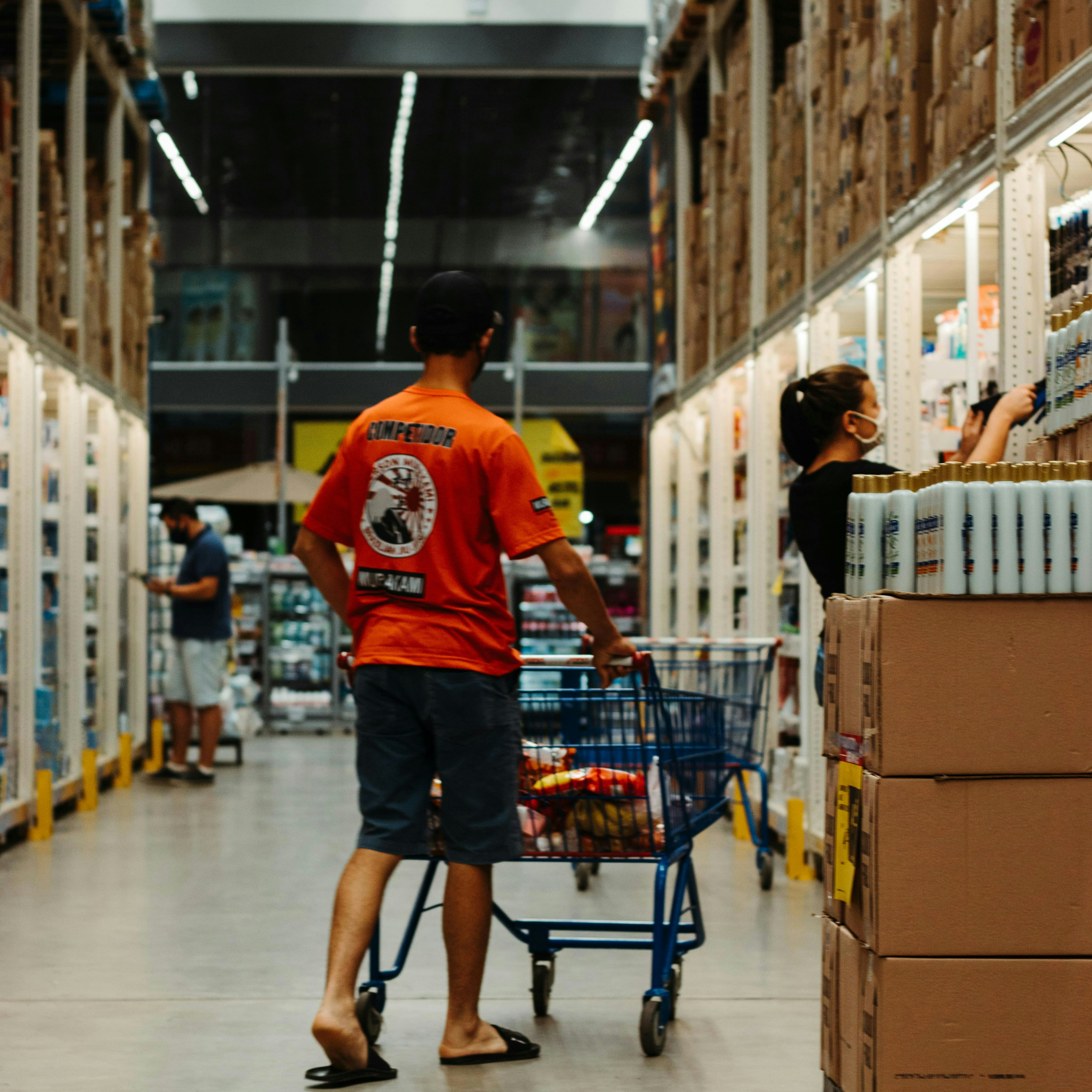 Un client pousse un chariot dans l'allée d'un magasin de type entrepôt, où les rayons de chaque côté sont chargés de marchandises empilées.