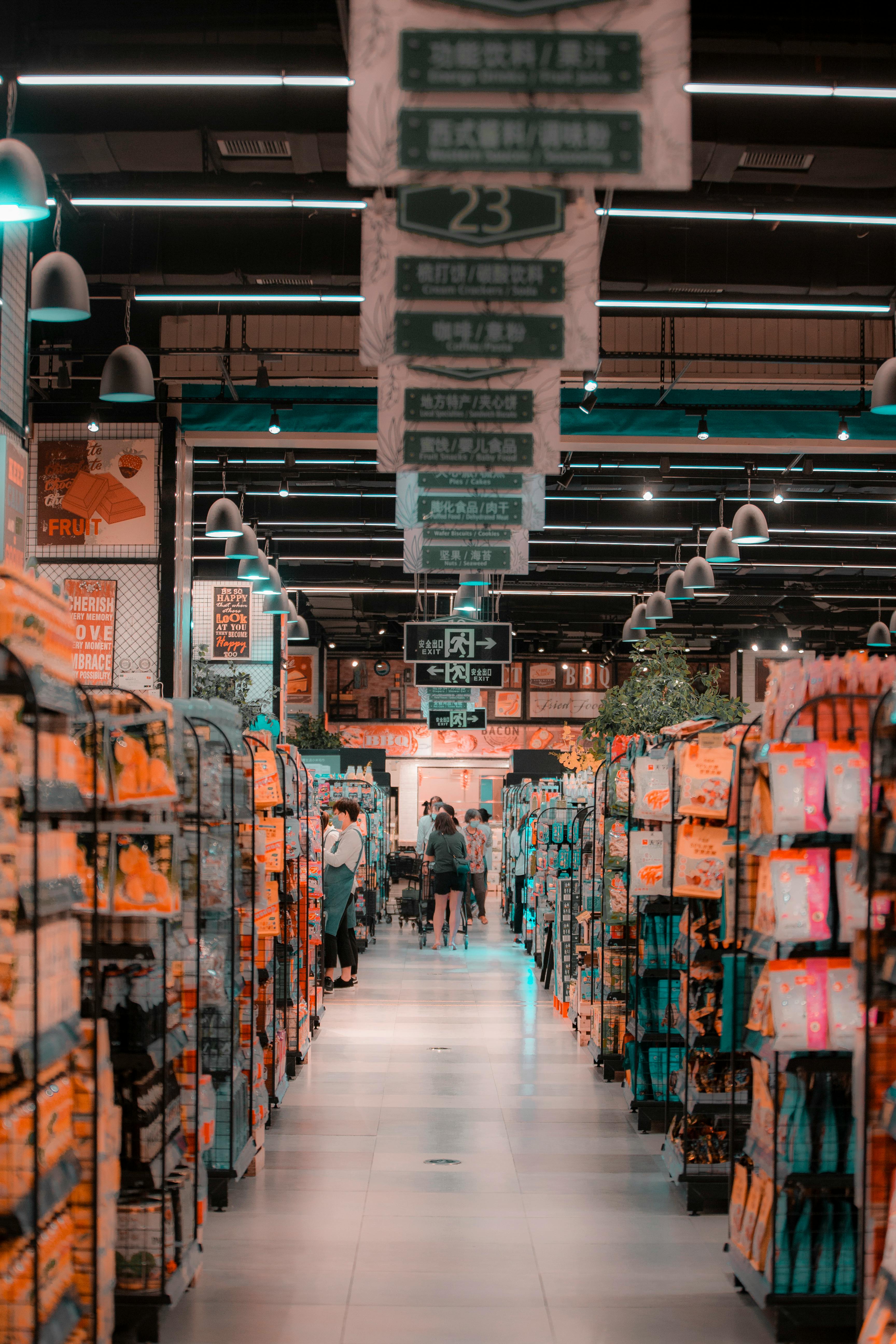 Les clients parcourent une longue allée de supermarché bordée de rayons bien garnis, au-dessus desquels sont suspendus des panneaux verts indiquant les catégories en chinois.