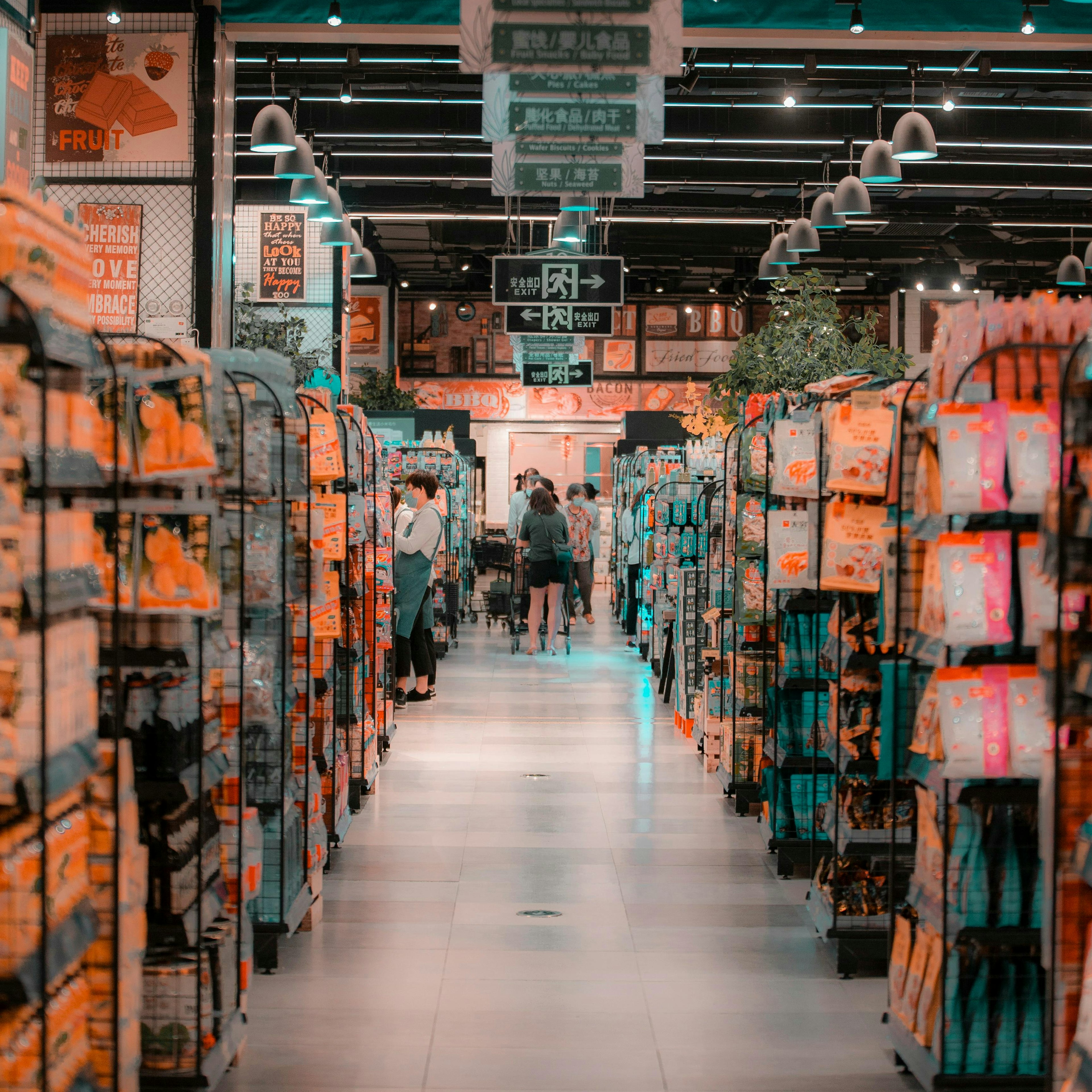 Les clients parcourent une longue allée de supermarché bordée de rayons bien garnis, au-dessus desquels sont suspendus des panneaux verts indiquant les catégories en chinois.