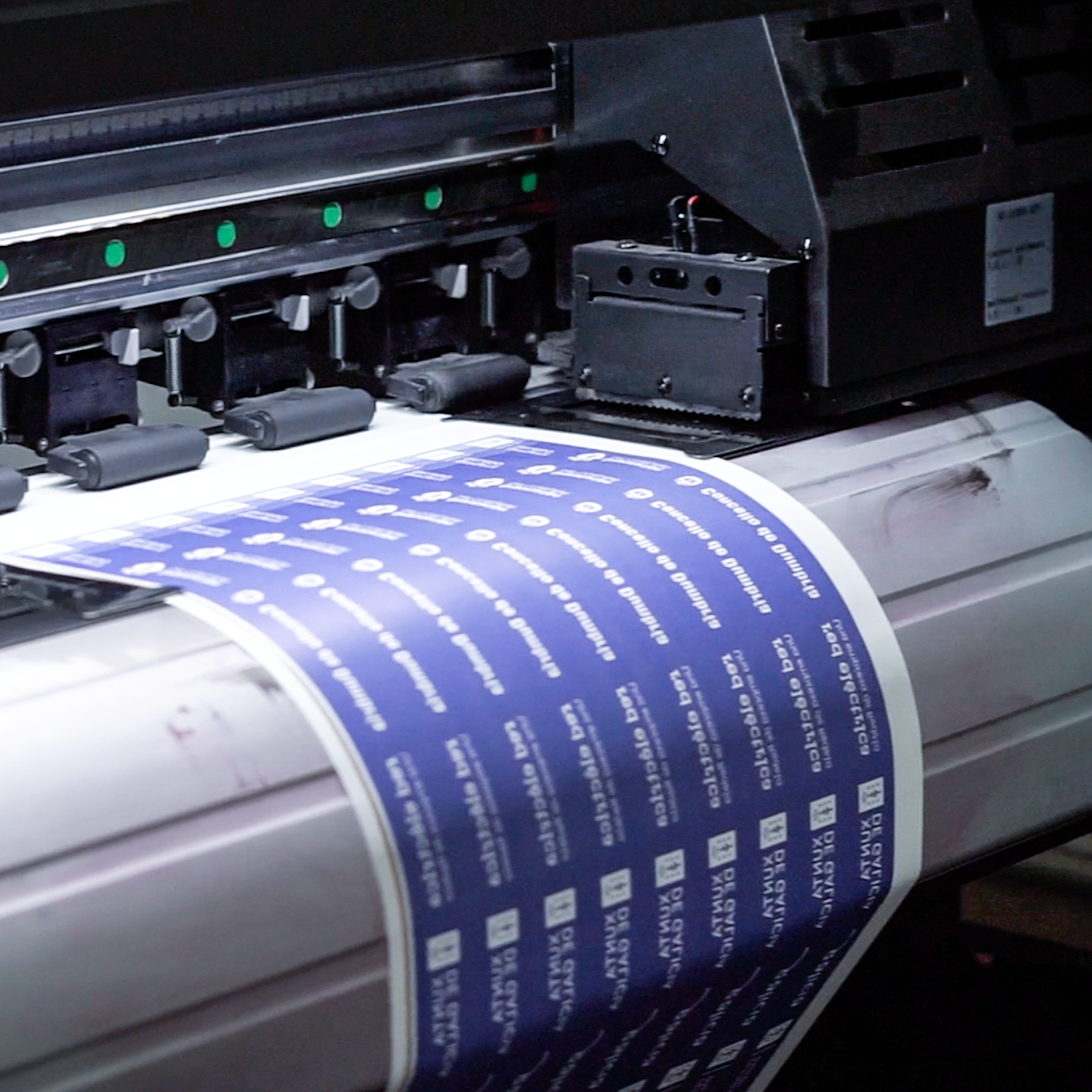 A wide-format printer feeds a roll of material printing a repeating blue design across the sheet.