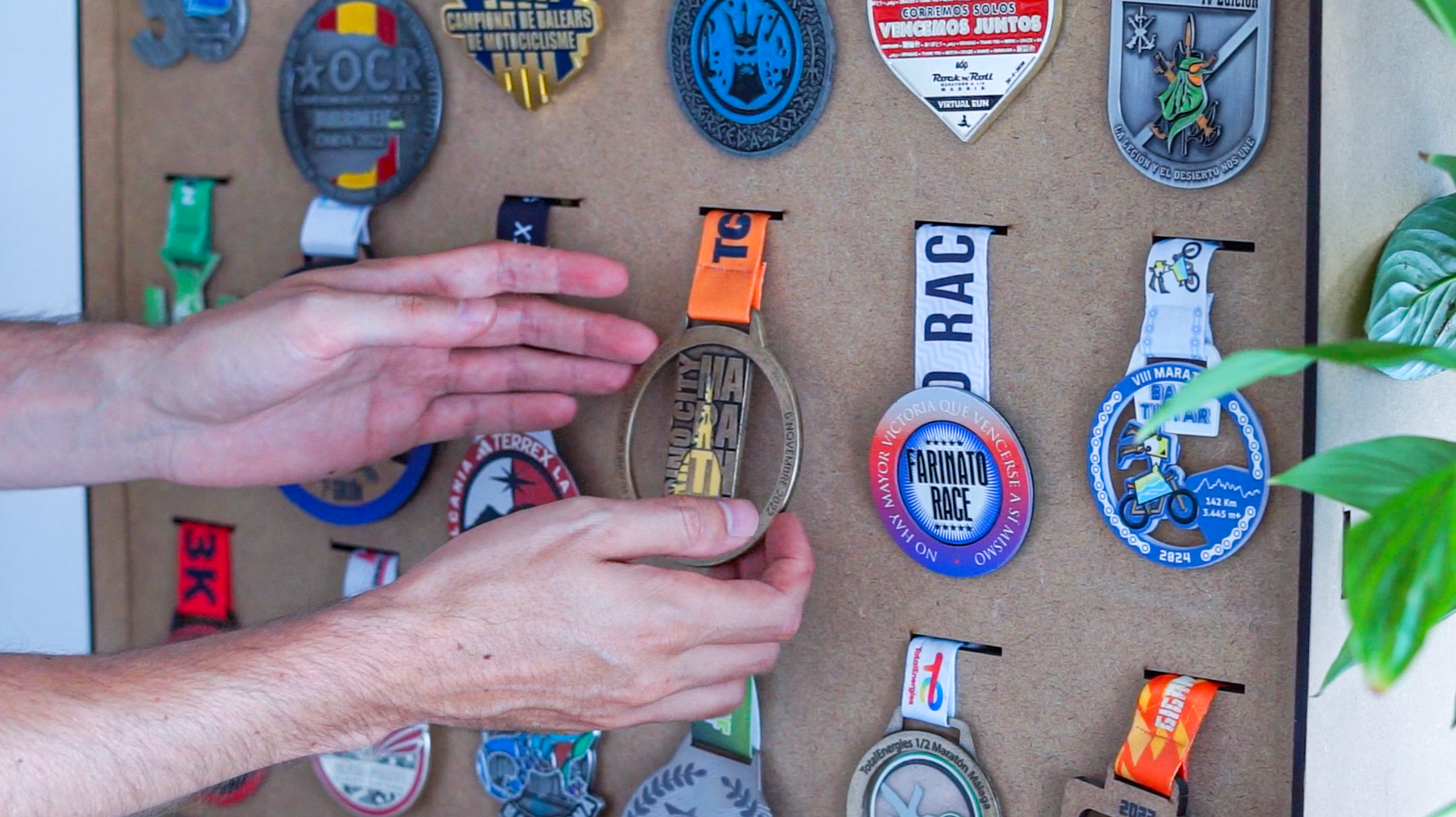 Two hands place a finished medal onto a wall rack already hung with a collection of race and event medals.
