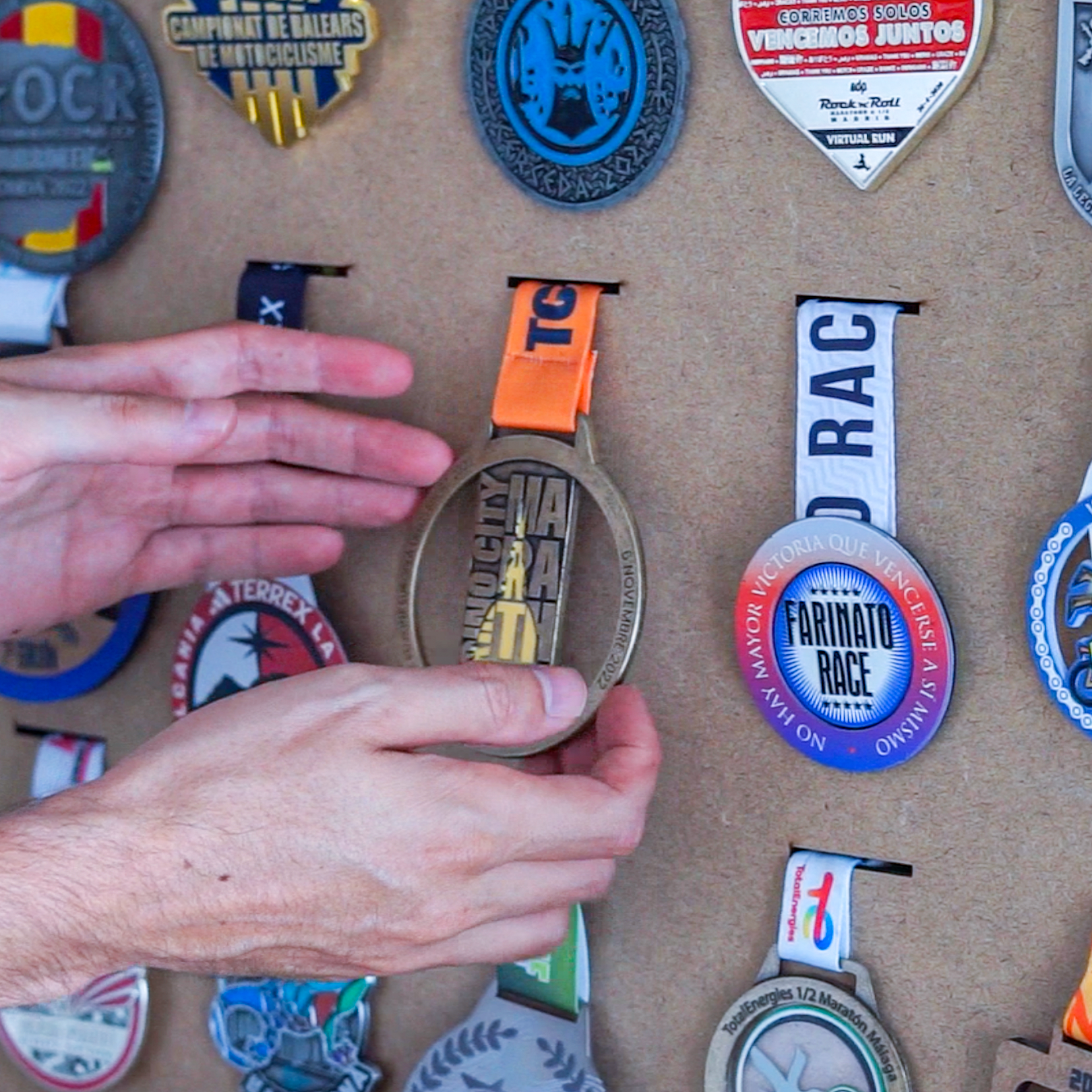 Two hands place a finished medal onto a wall rack already hung with a collection of race and event medals.