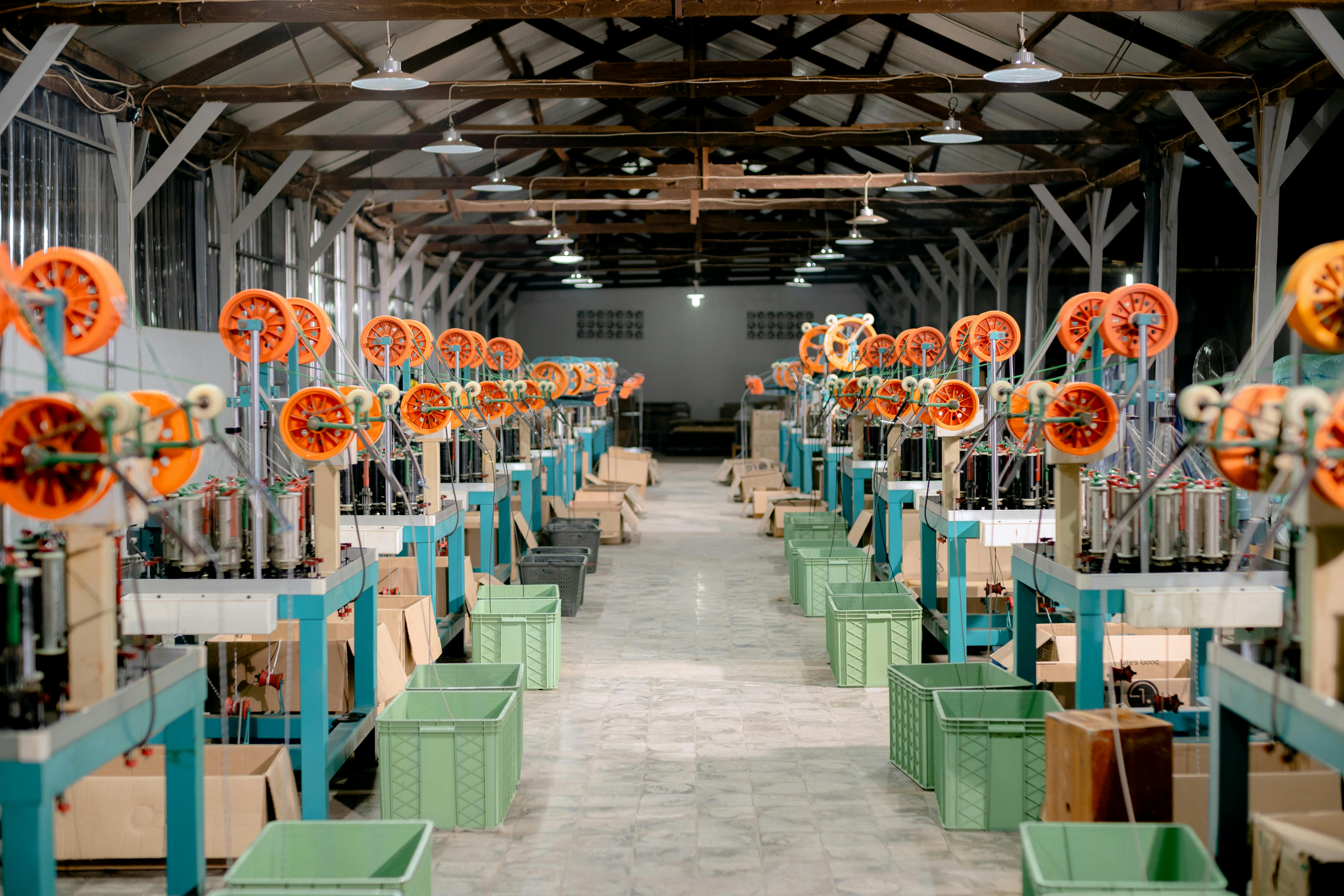 Deux longues rangées de machines à tresser, équipées de bobines orange et de bacs de collecte verts, s'étendent sur toute la longueur du hall d'une usine textile.