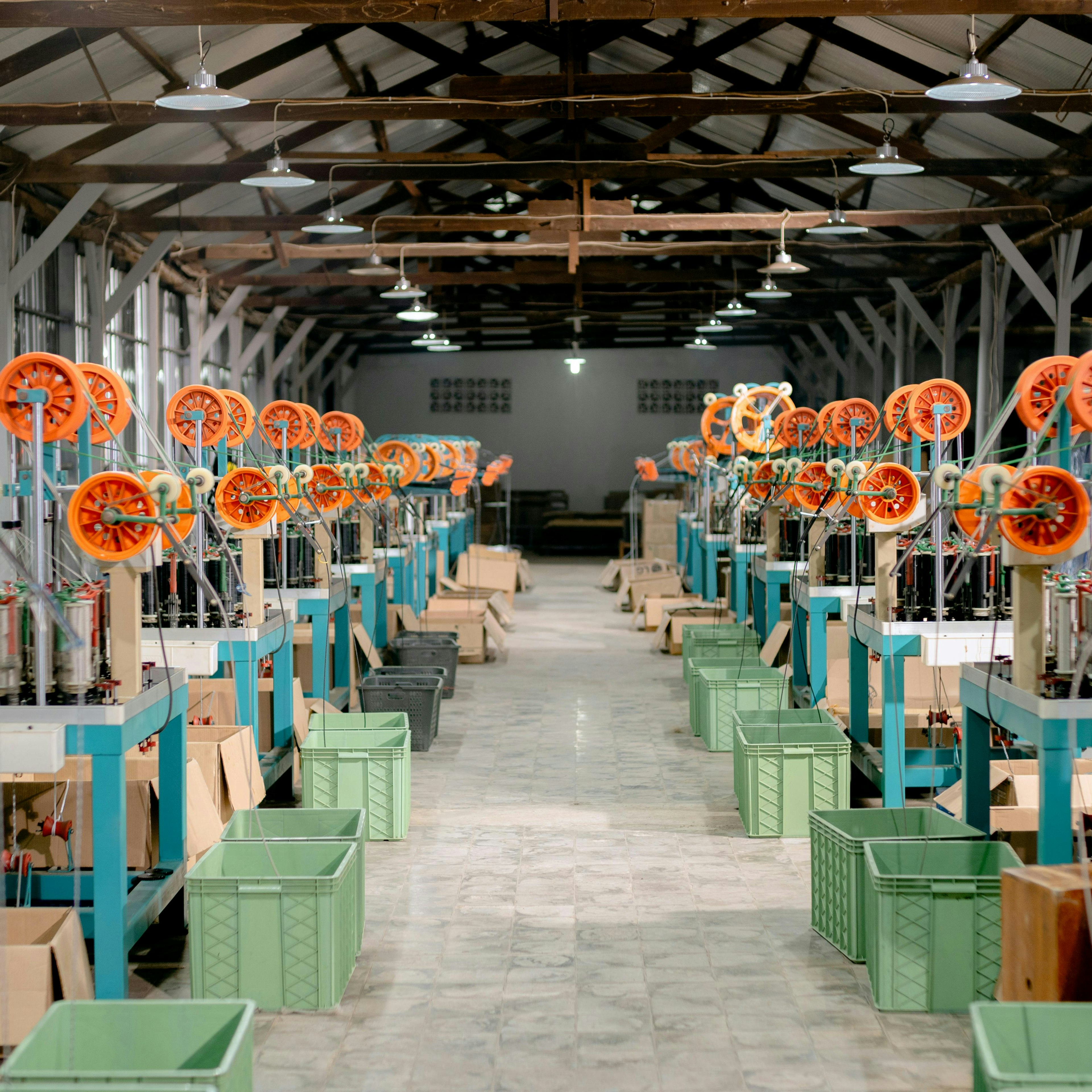 Deux longues rangées de machines à tresser, équipées de bobines orange et de bacs de collecte verts, s'étendent sur toute la longueur du hall d'une usine textile.