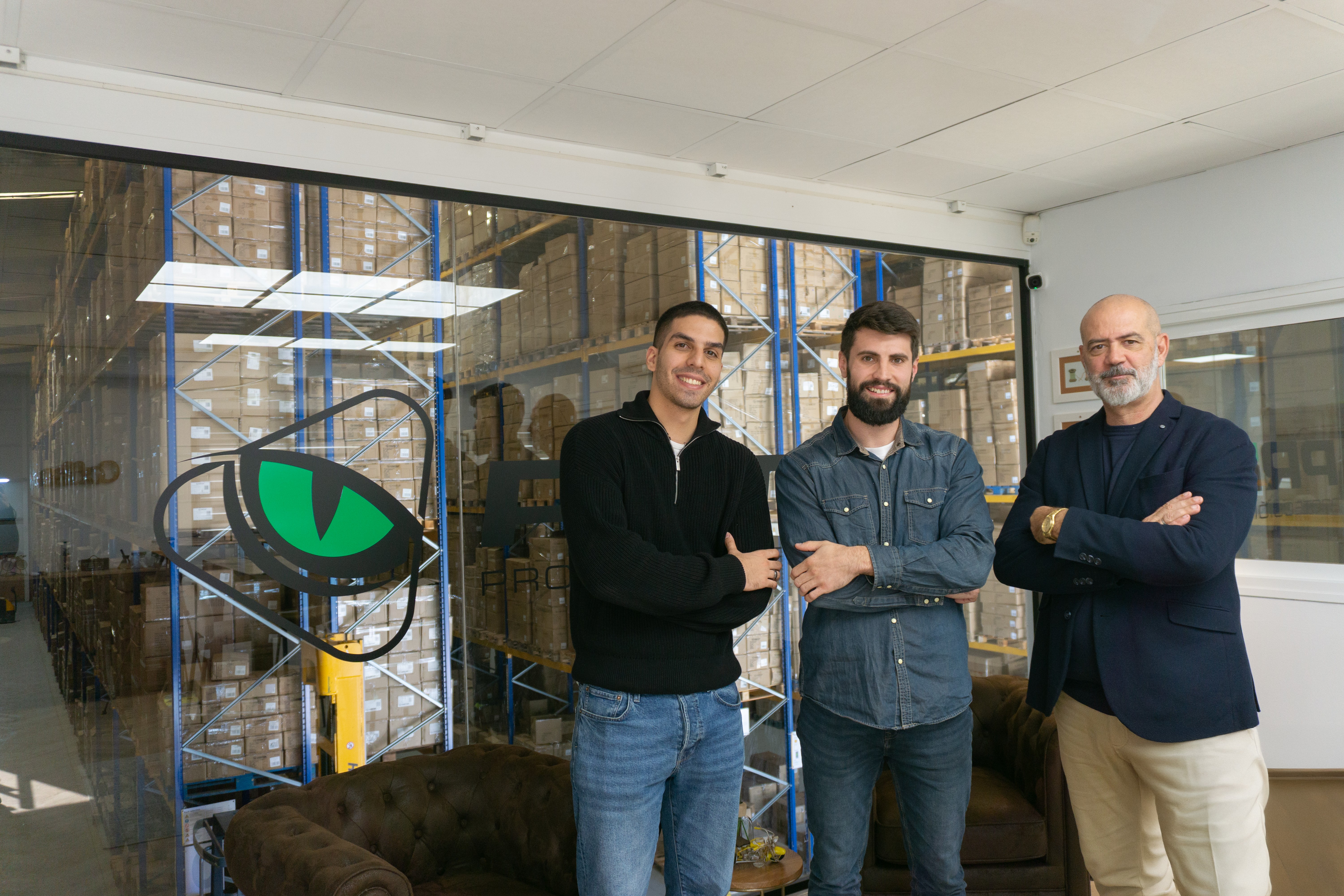 Three members of the Prima team stand with arms crossed in front of a glass wall bearing the company's eye logo, the stocked warehouse visible behind.