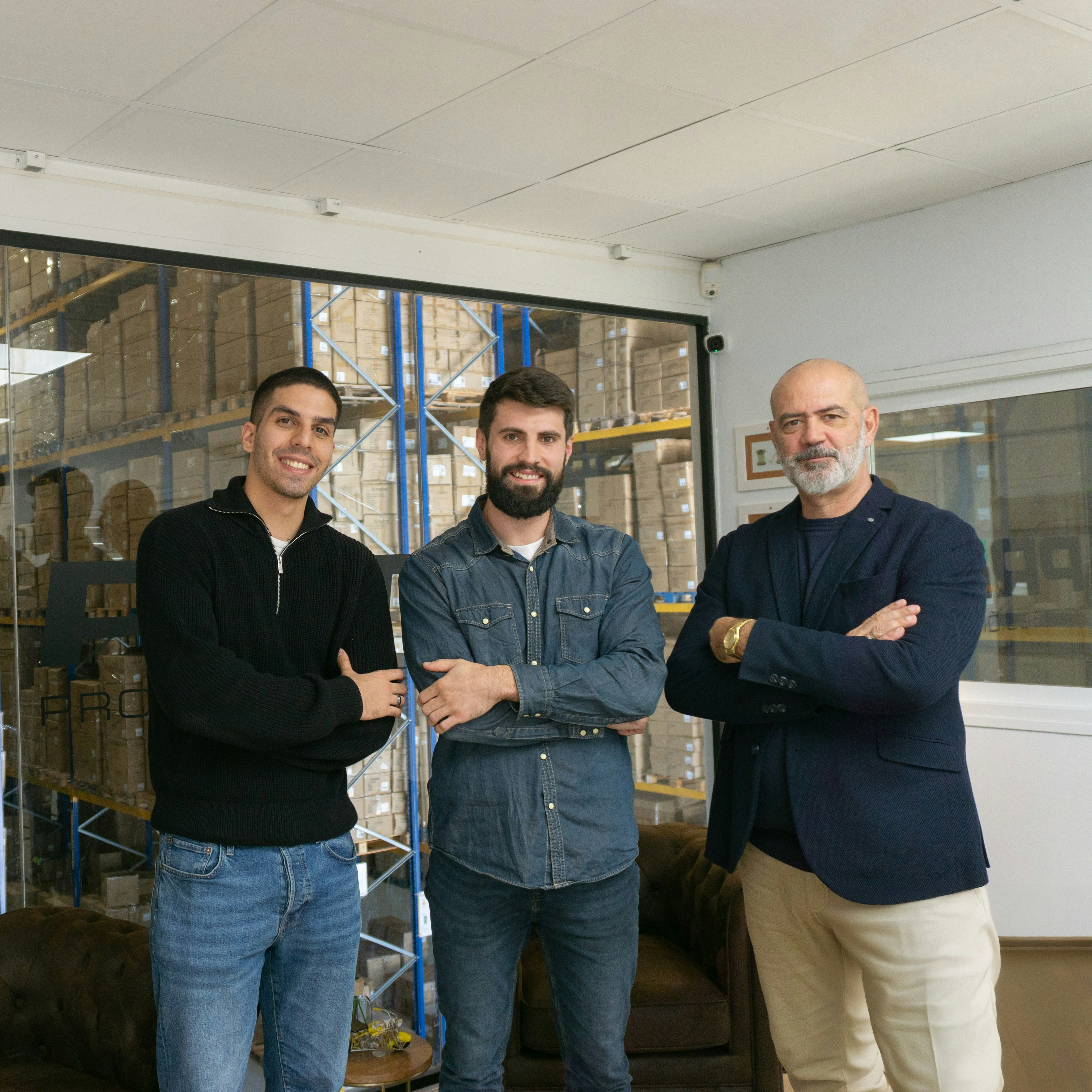 Three members of the Prima team stand with arms crossed in front of a glass wall bearing the company's eye logo, the stocked warehouse visible behind.