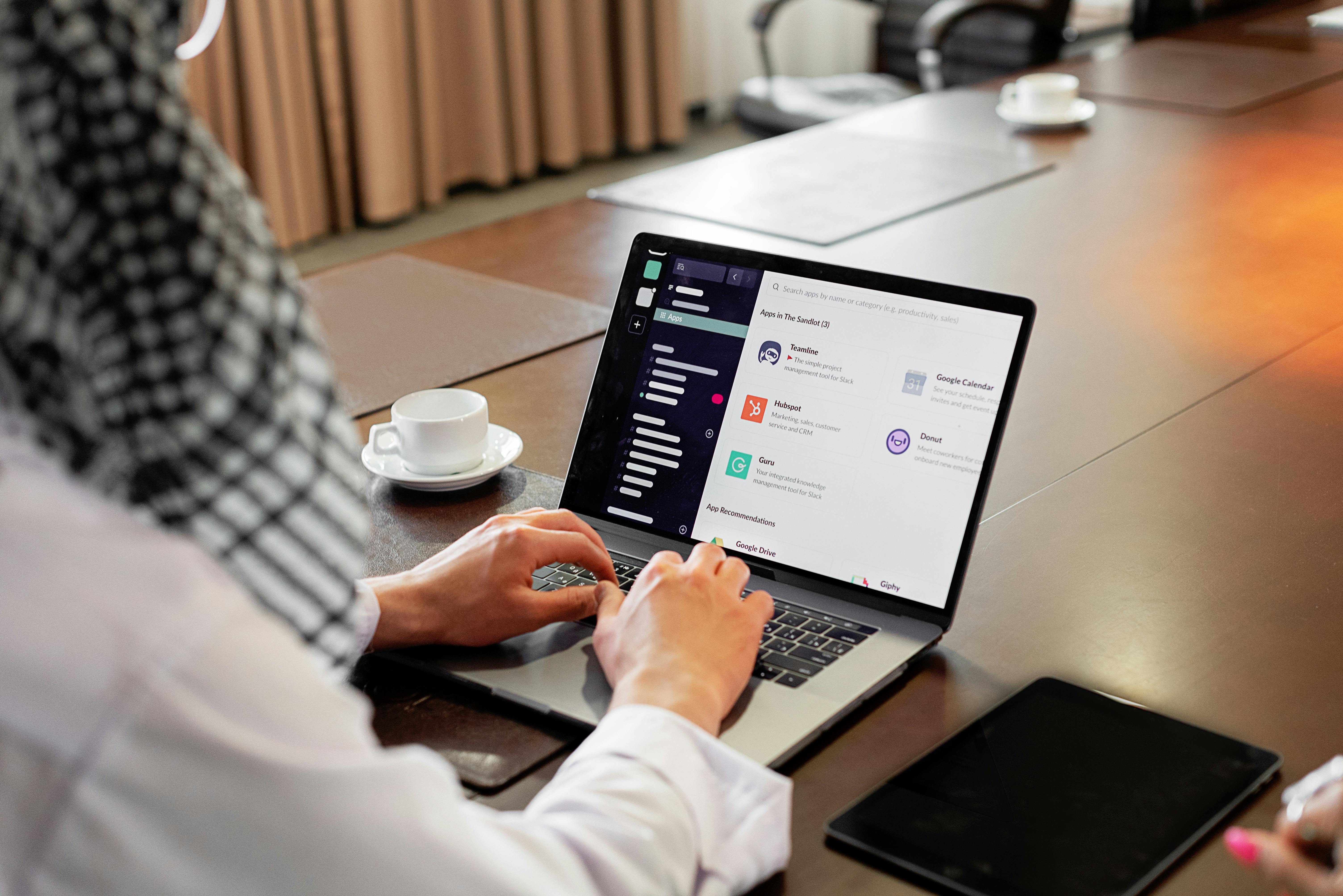 Over-the-shoulder view of someone using the Slack app directory on a laptop in a meeting room, with a coffee cup beside them.