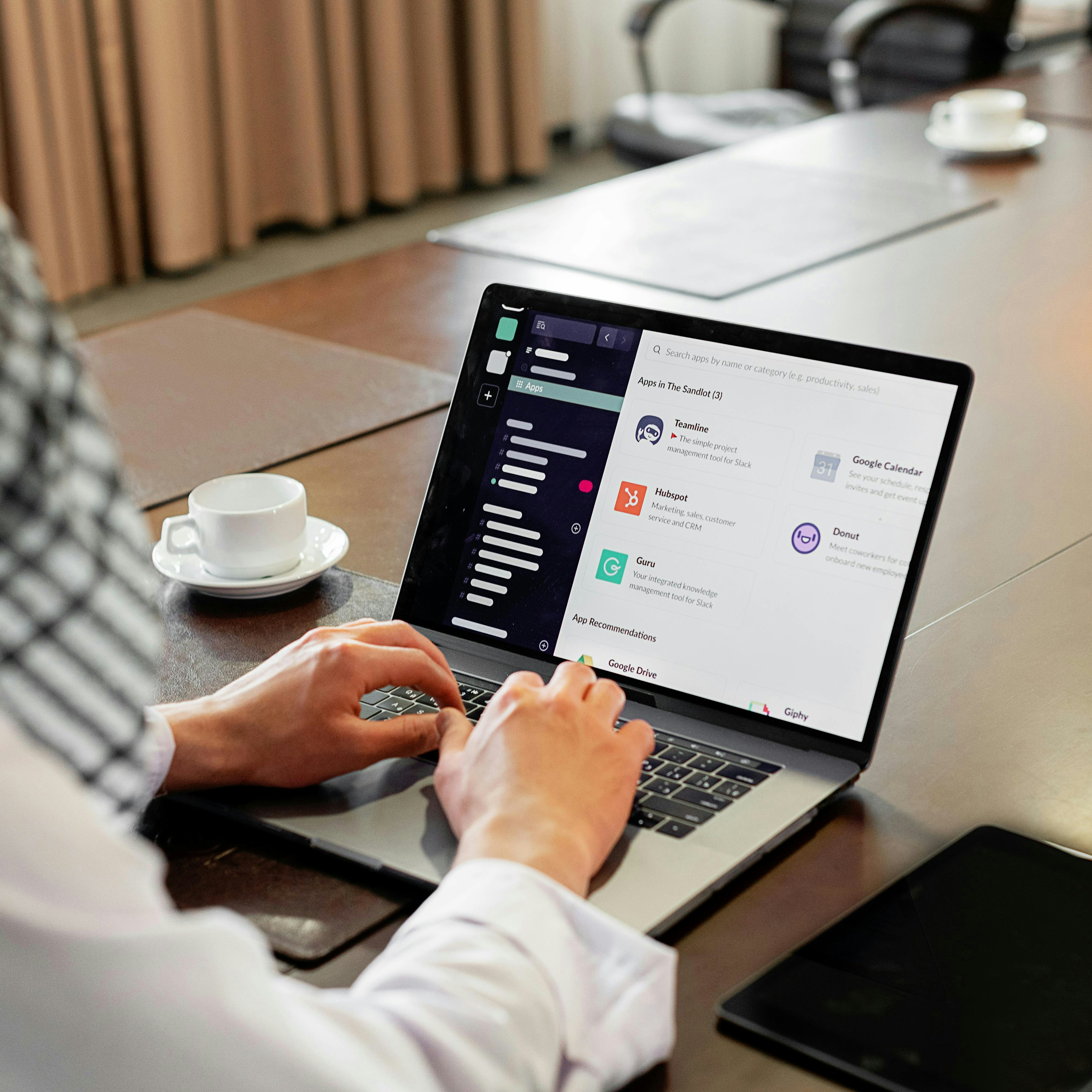 Vue par-dessus l'épaule d'une personne utilisant le répertoire d'applications Slack sur un ordinateur portable dans une salle de réunion, avec une tasse de café à côté d'elle.