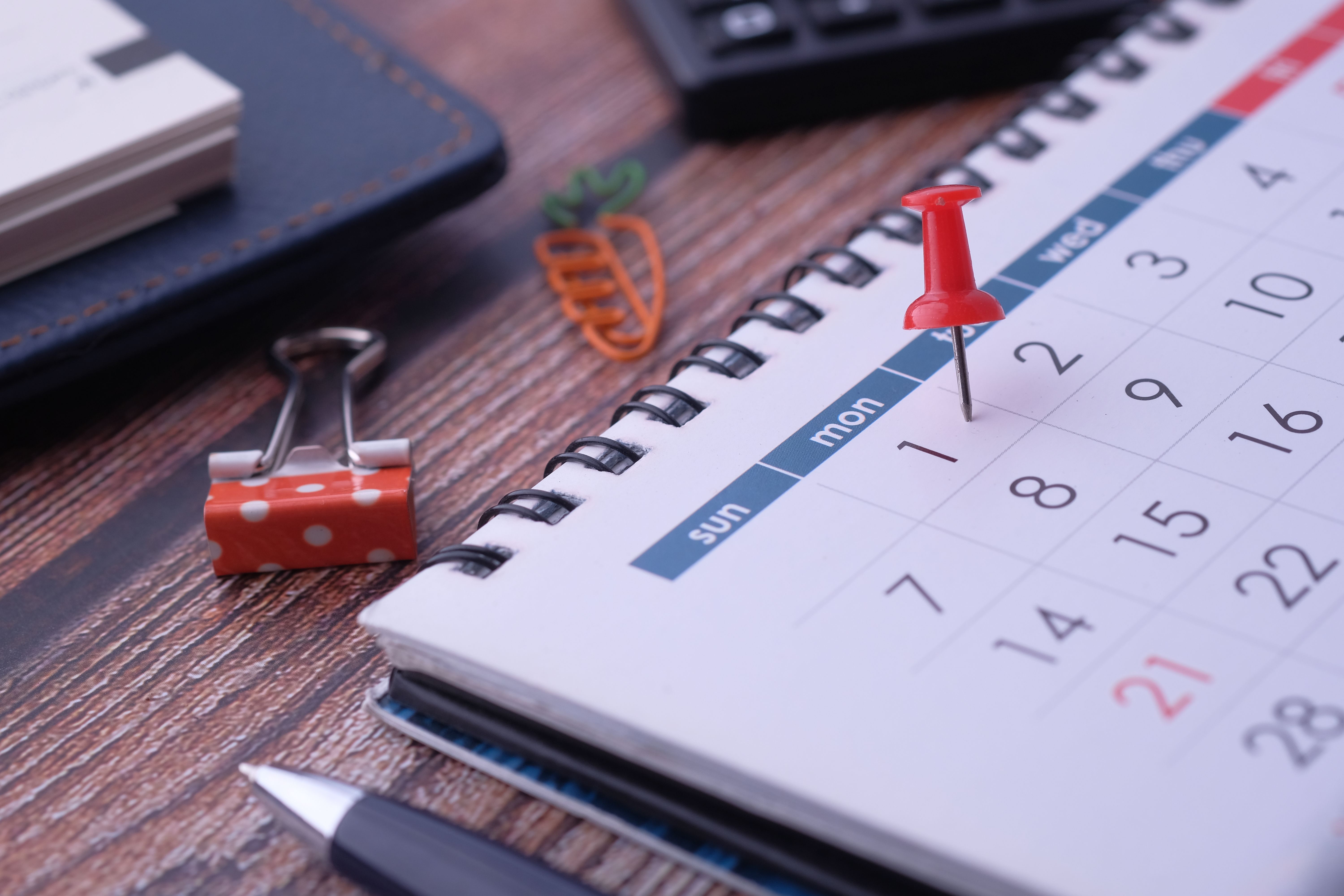 Calendario de planificación con una chincheta roja marcando una fecha, rodeado de elementos de oficina en un escritorio.