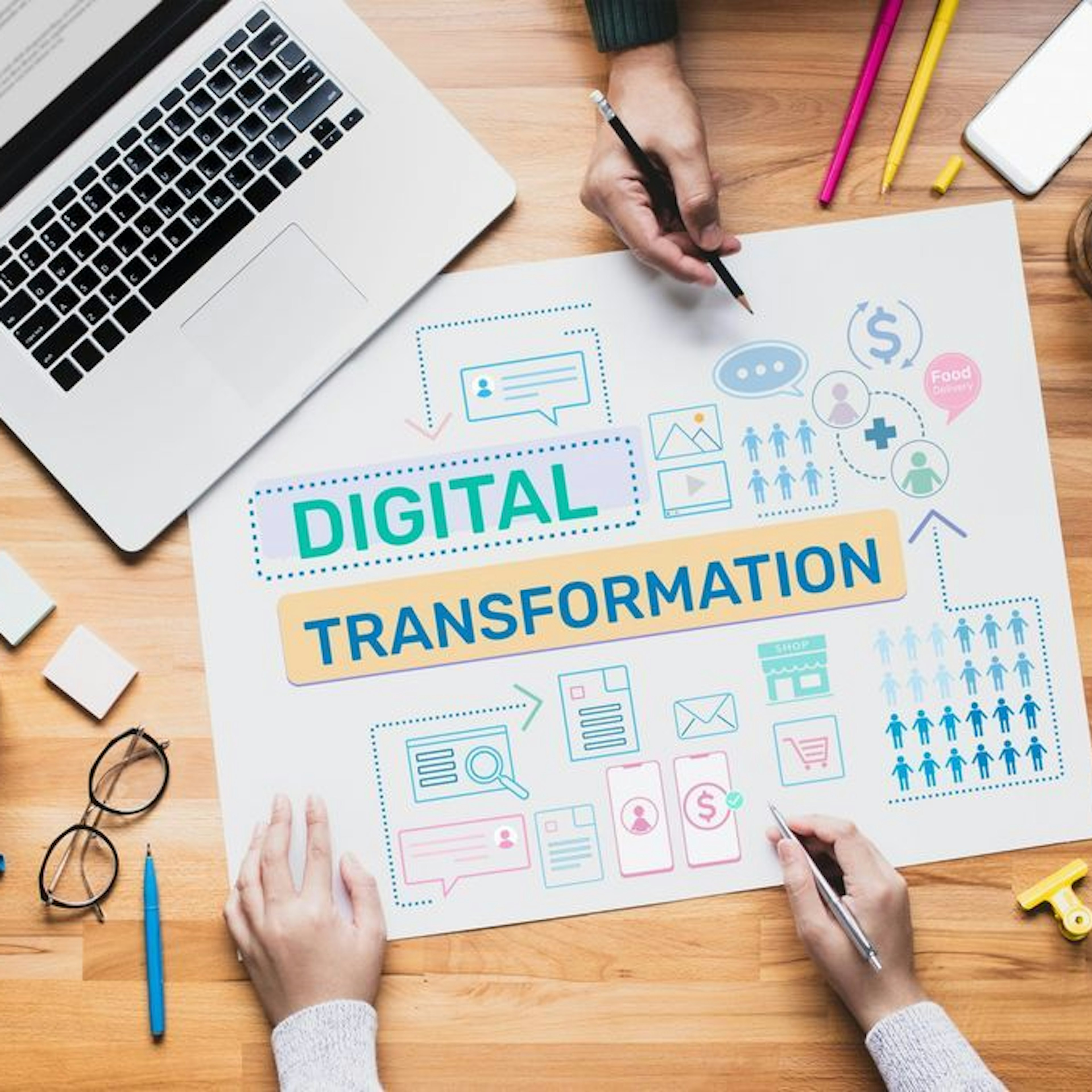 Two people working on a digital transformation project, surrounded by diagrams, icons, and office supplies on a wooden desk.