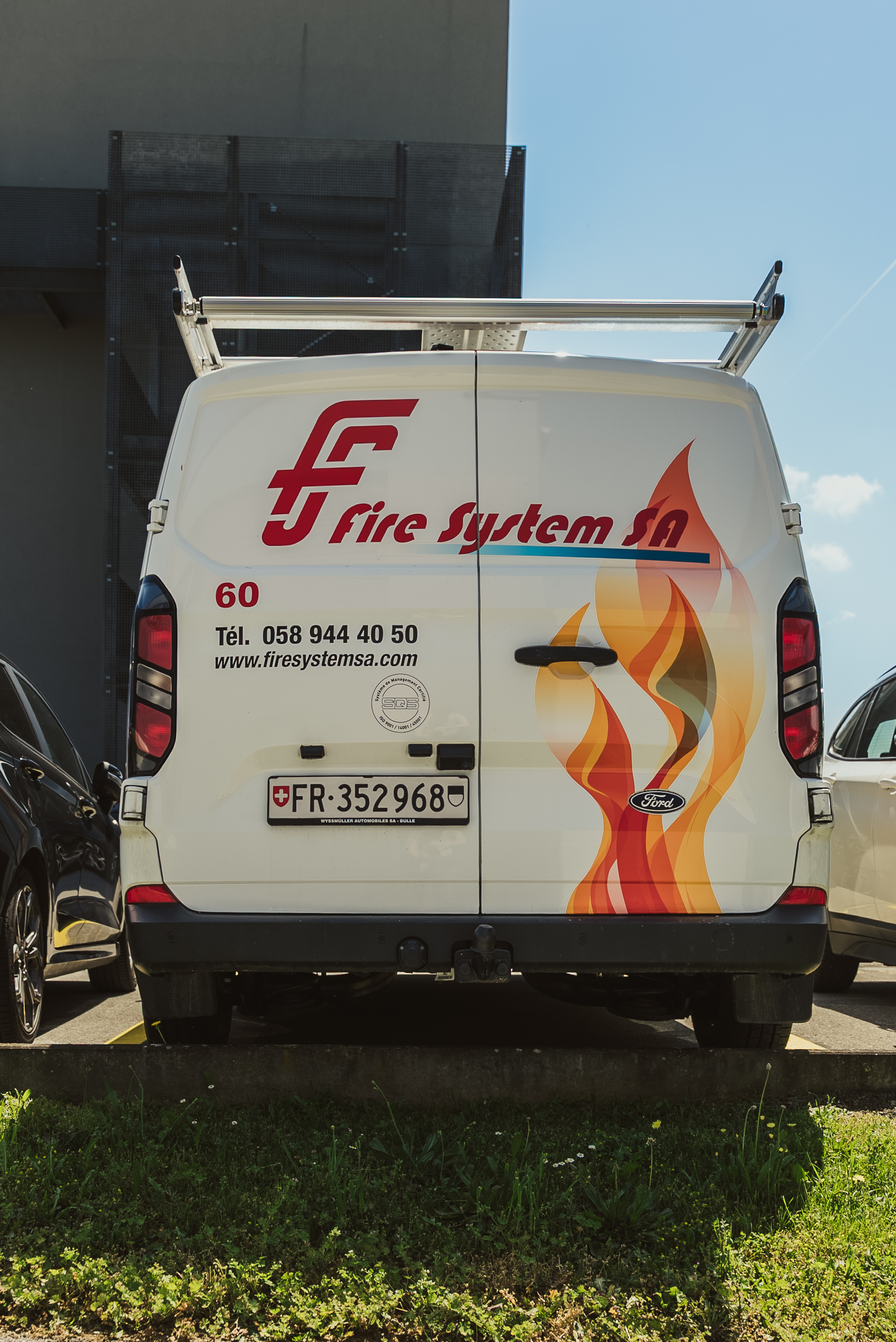 The rear of a white Fire System SA Ford van with the company logo, flame graphic and contact details, parked outside the premises.
