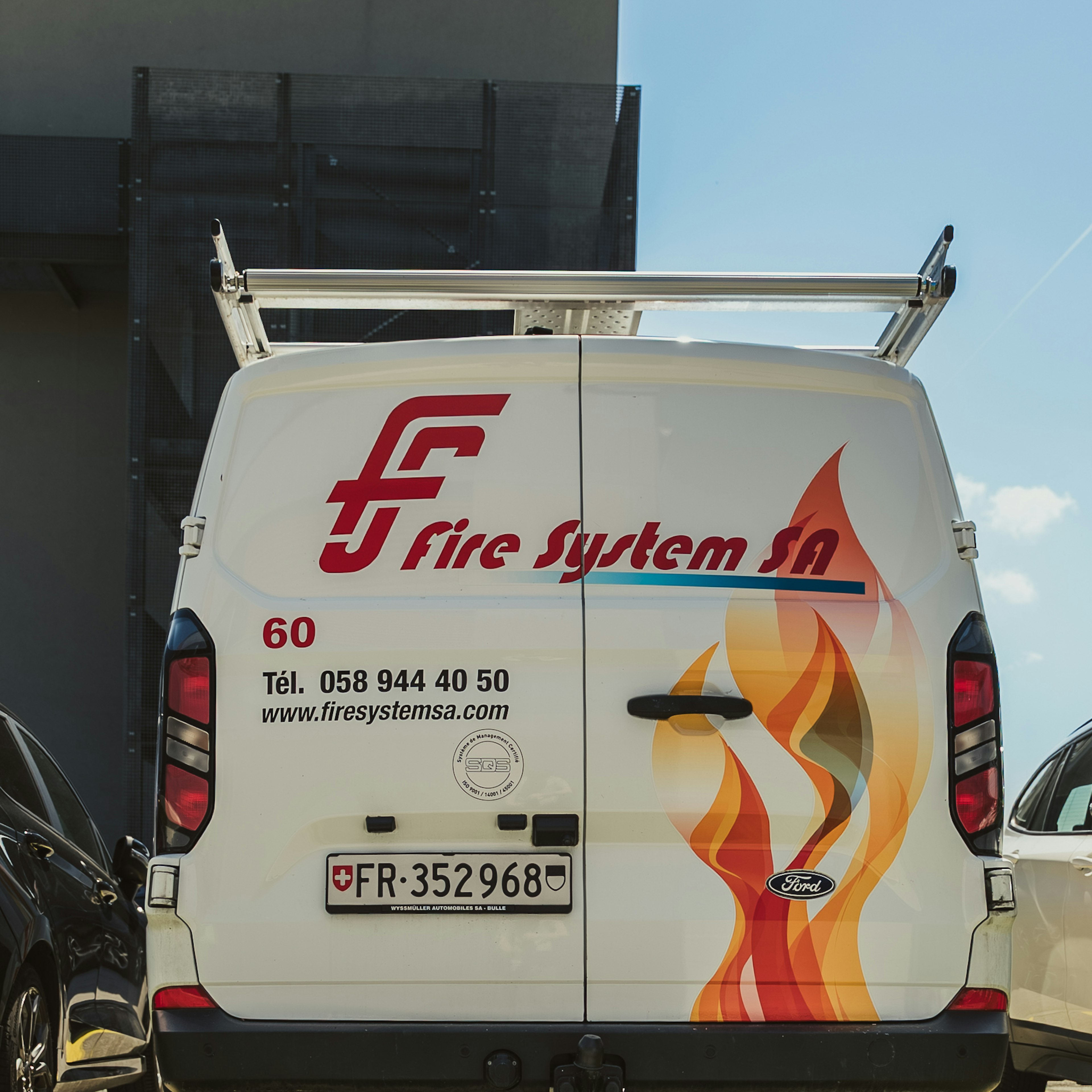 L'arrière d'une camionnette Ford blanche de la société Fire System SA, arborant le logo de l'entreprise, un motif de flammes et les coordonnées, garée devant les locaux.