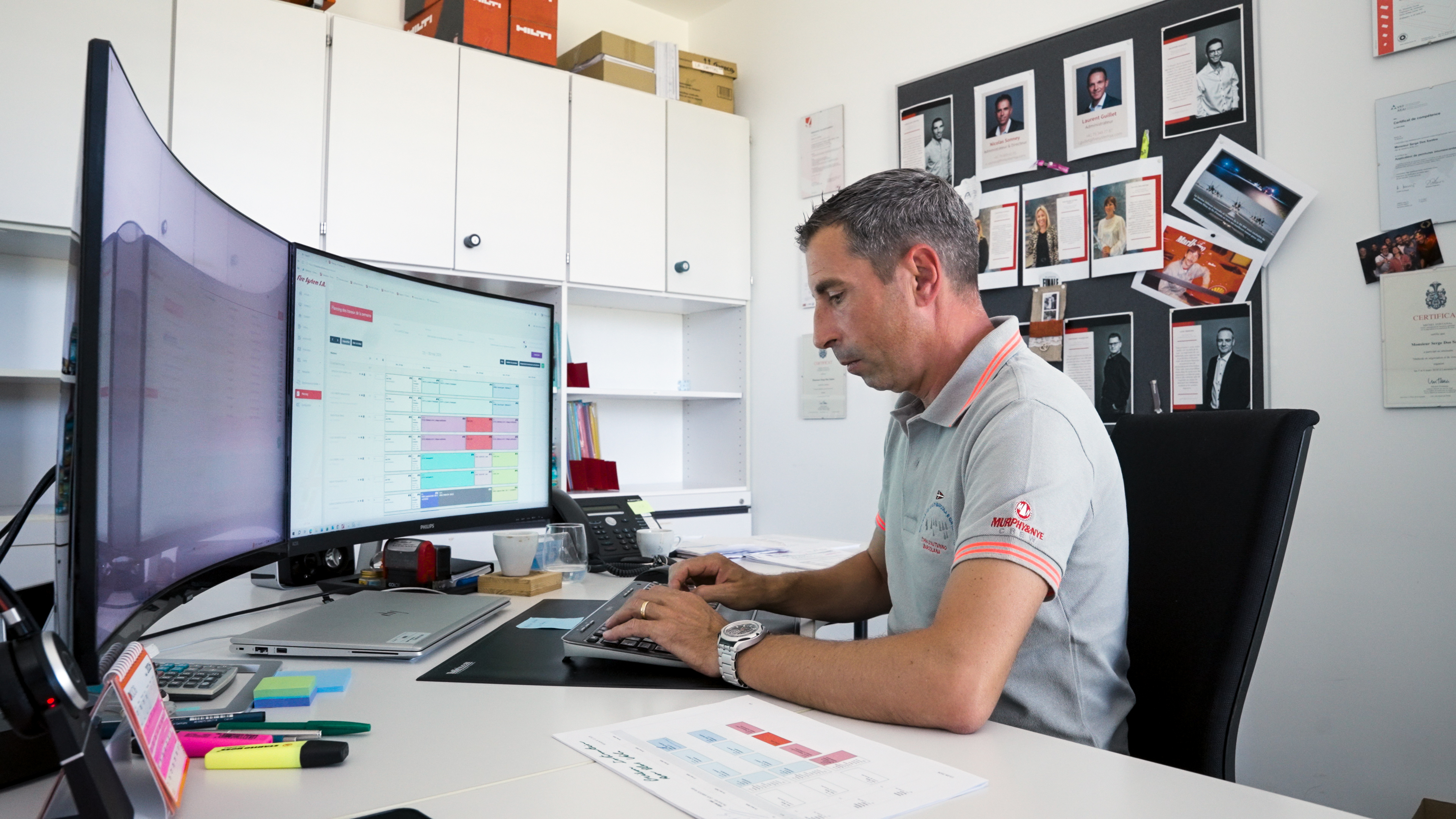 A man in a Fire System polo shirt works at a dual-monitor desk, reviewing the FS Gestion scheduling tool on a curved screen.