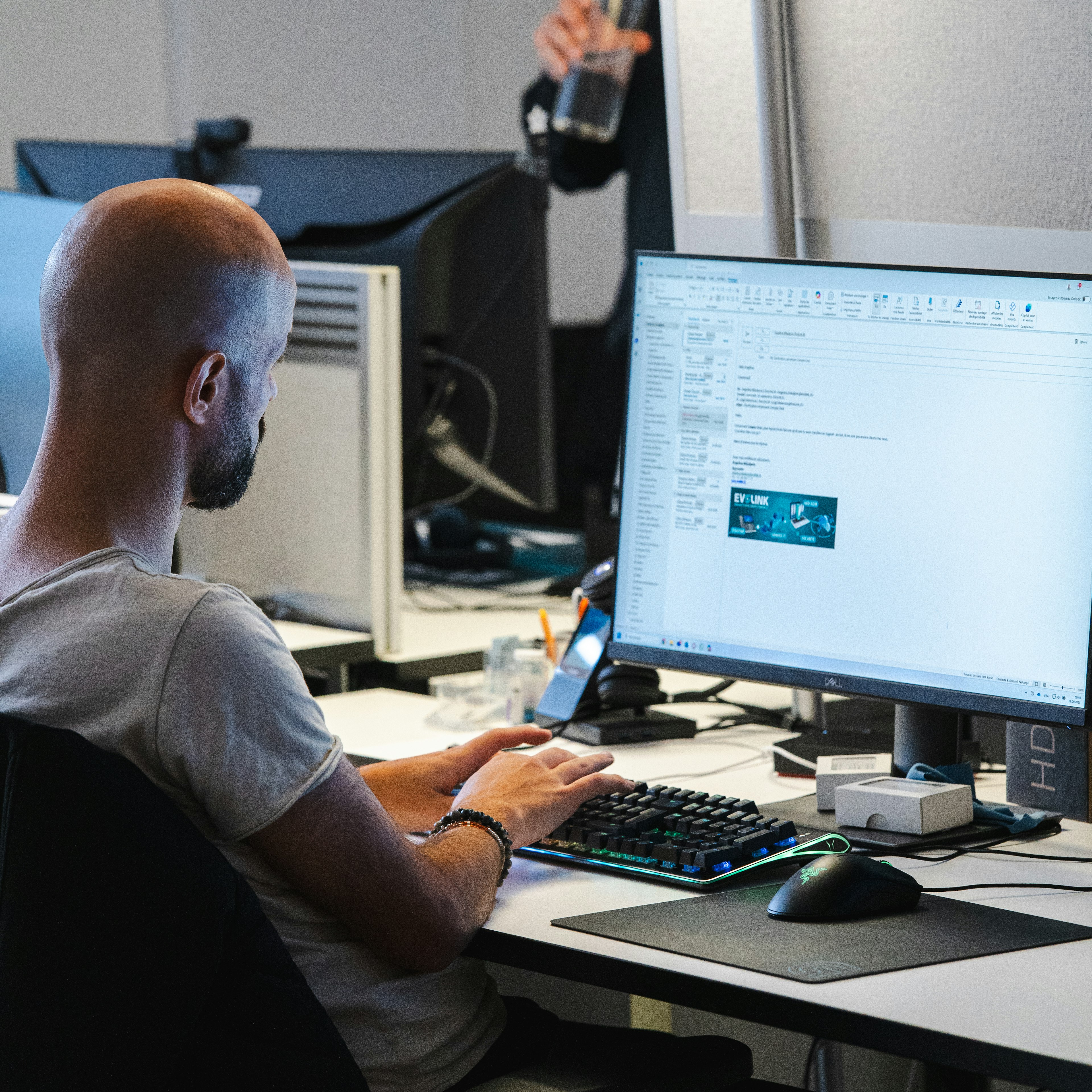Un employé d'Evolink travaille devant son ordinateur au bureau ; on aperçoit le logo d'Evolink dans un e-mail ouvert, avec un collègue en arrière-plan.