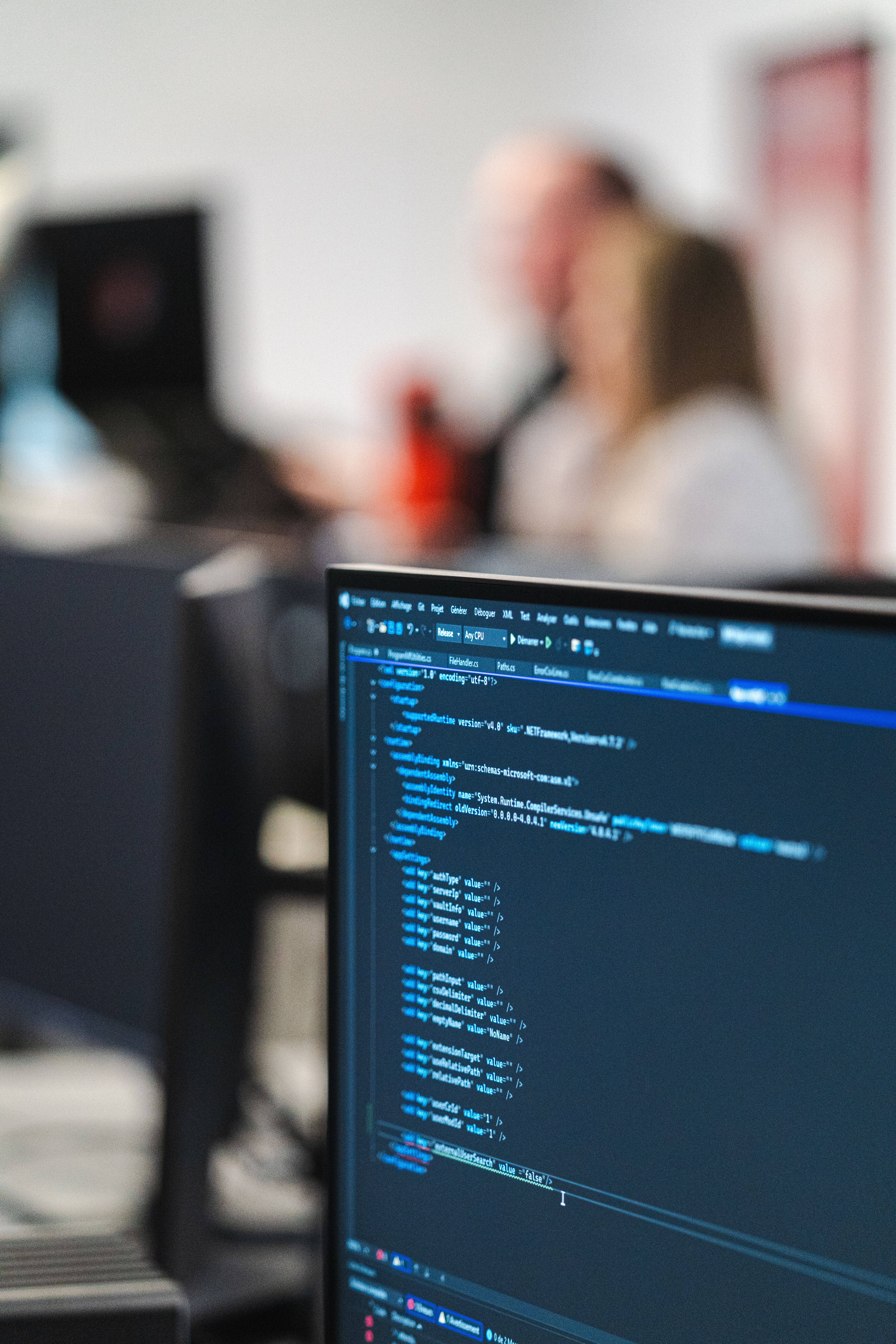 A monitor displays lines of code in a development environment, with colleagues talking in the blurred background.