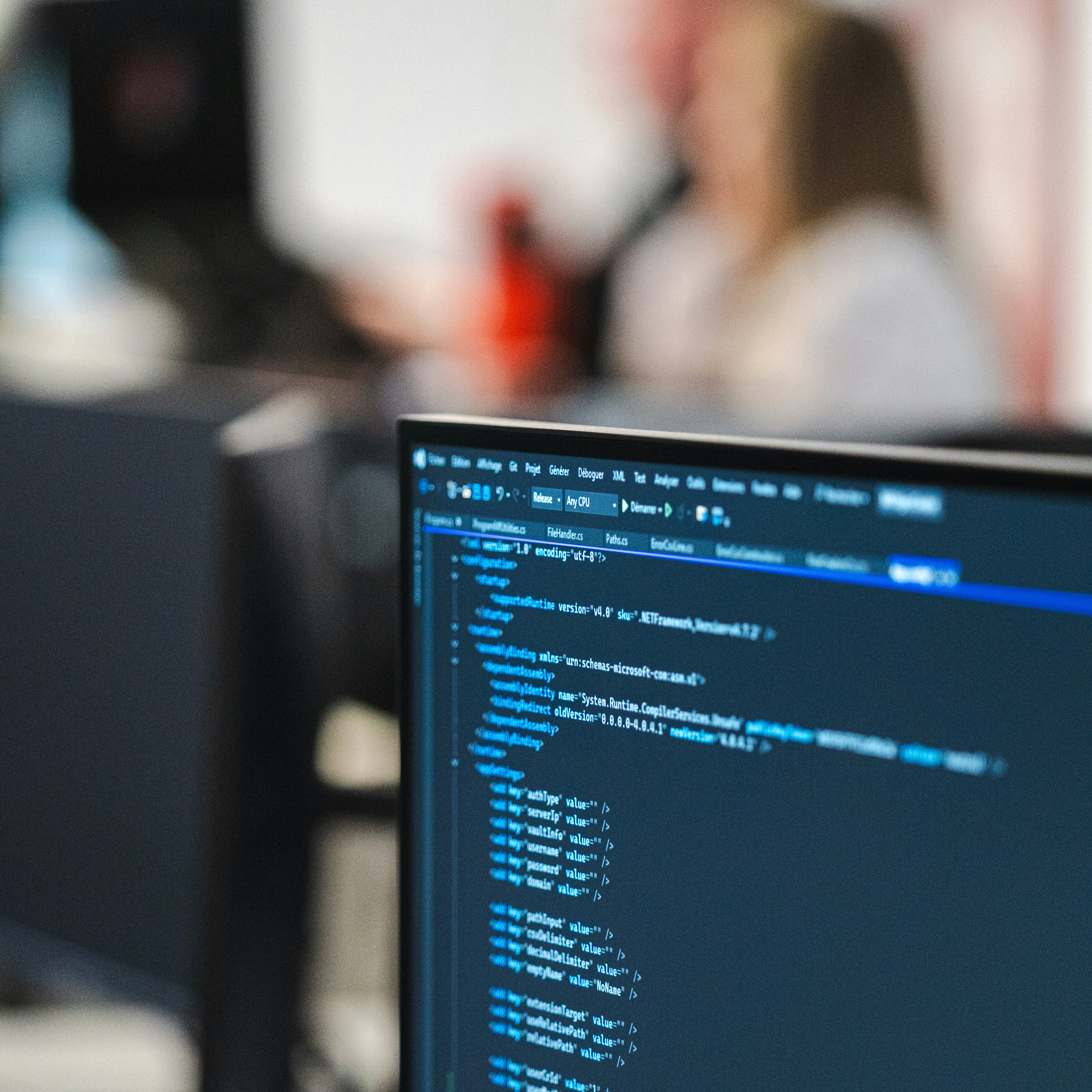 Un écran affiche des lignes de code dans un environnement de développement, tandis que des collègues discutent en arrière-plan, flous.