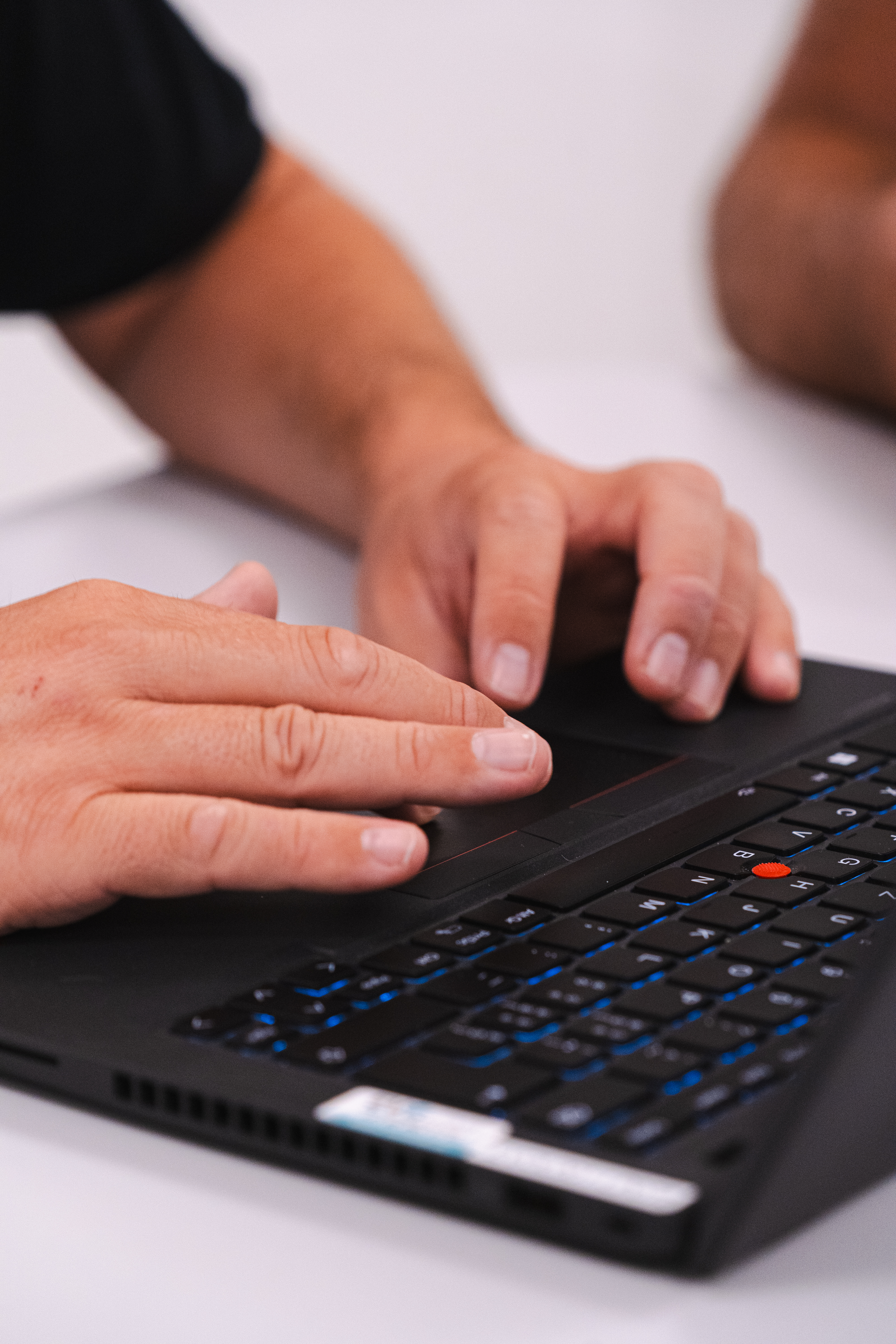Close-up of a person's hands typing on a backlit black laptop keyboard with a red trackpoint, against a white desk.