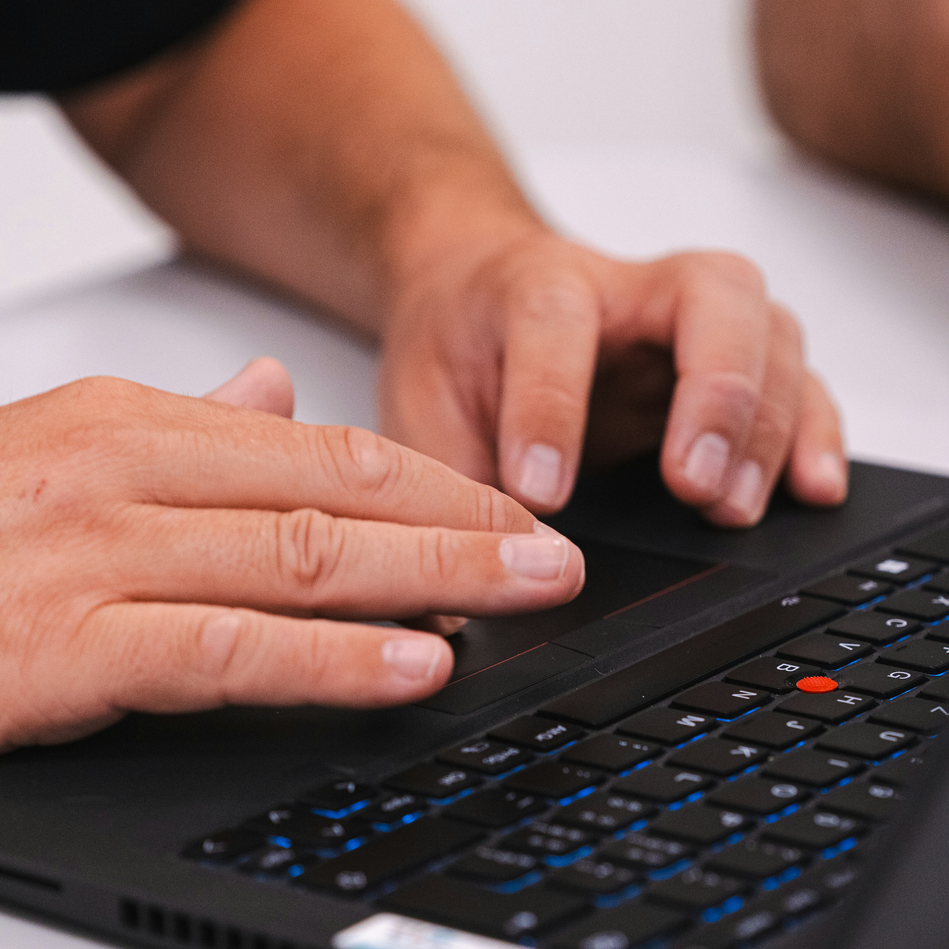 Gros plan sur les mains d'une personne tapant sur le clavier rétroéclairé d'un ordinateur portable noir équipé d'un trackpoint rouge, sur un bureau blanc.