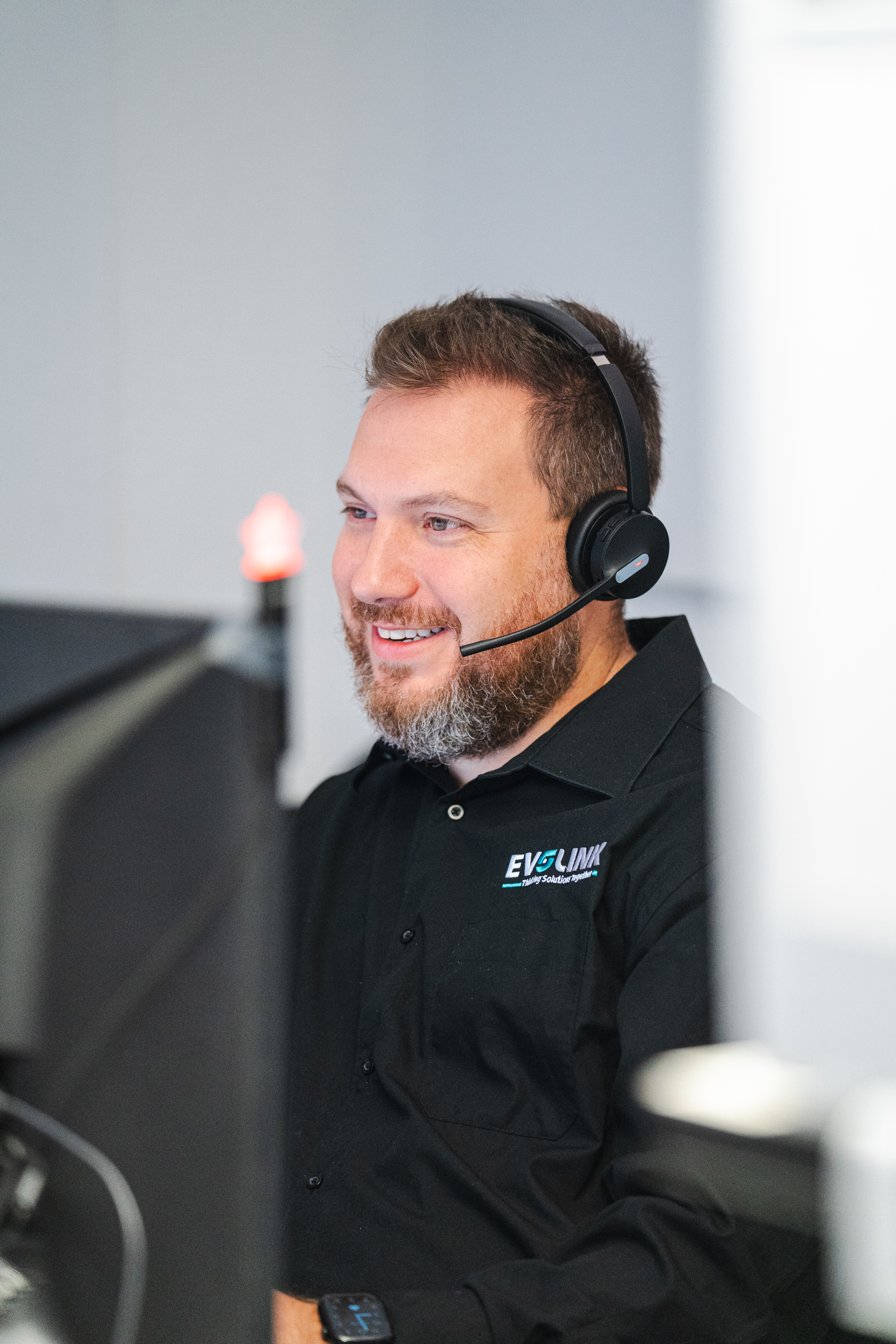 A smiling Evolink support agent in a branded black shirt wears a headset while working at his desk.