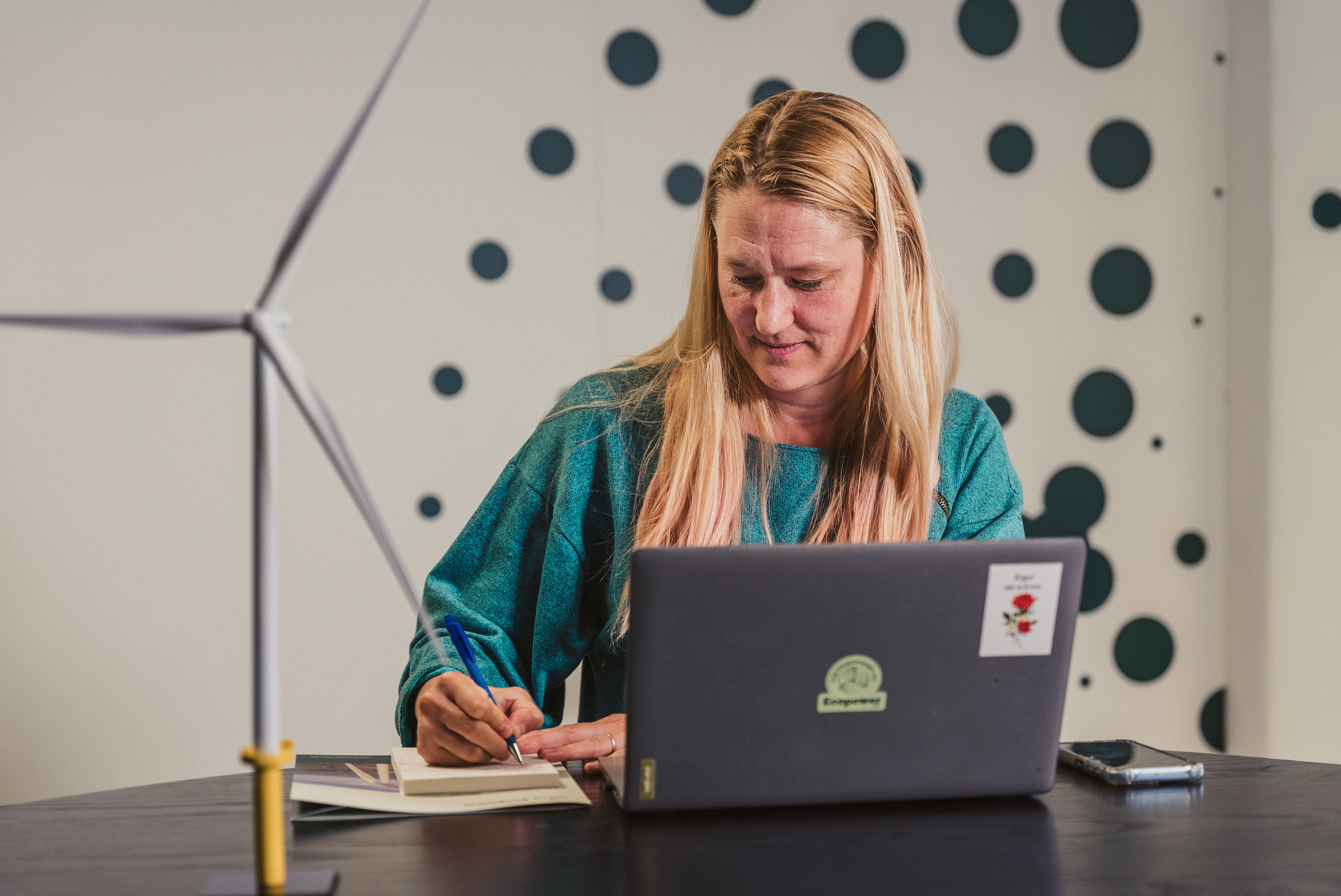 A woman in a teal top writes in a notebook beside a laptop with an Ecopower sticker, with a model wind turbine on the desk.