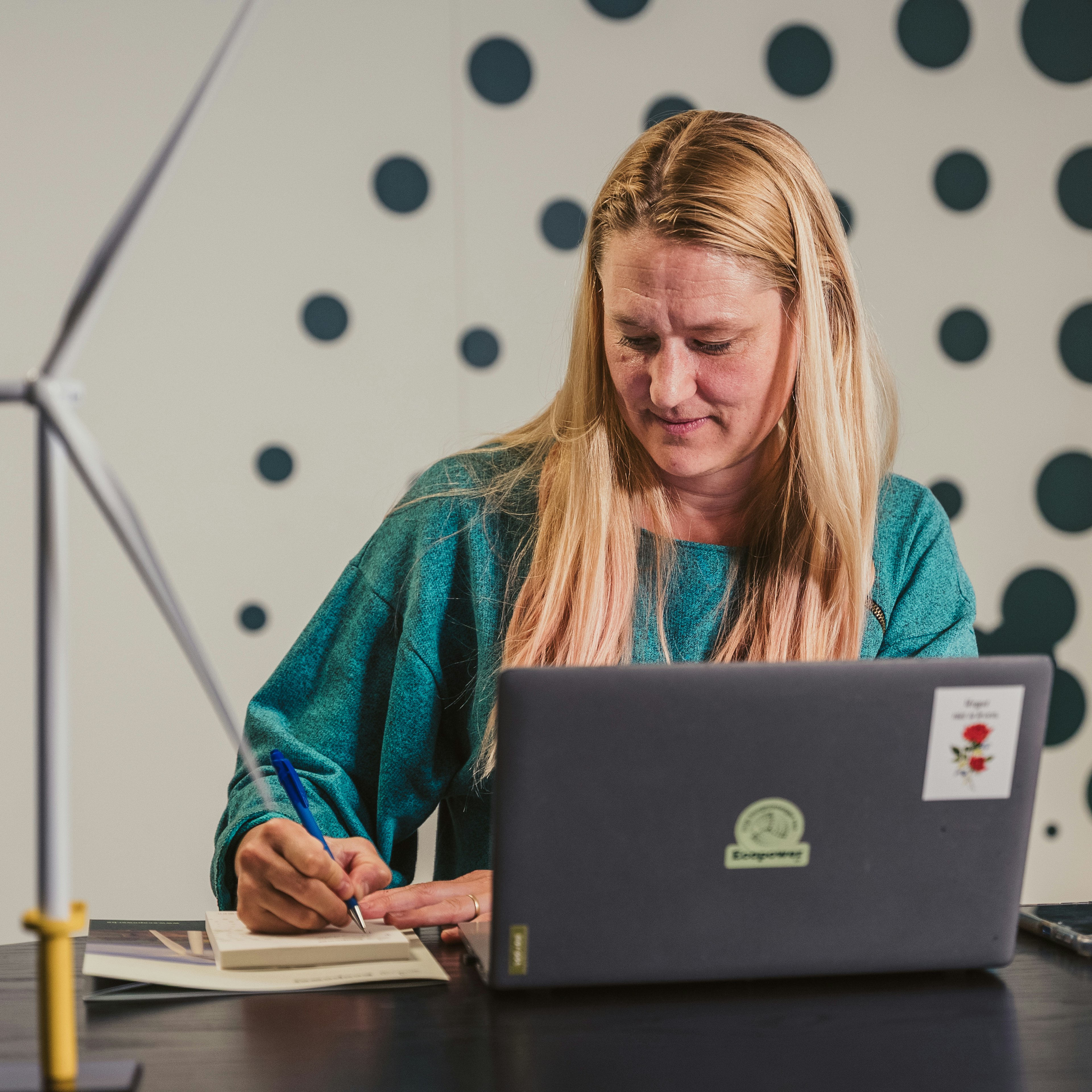 A woman in a teal top writes in a notebook beside a laptop with an Ecopower sticker, with a model wind turbine on the desk.